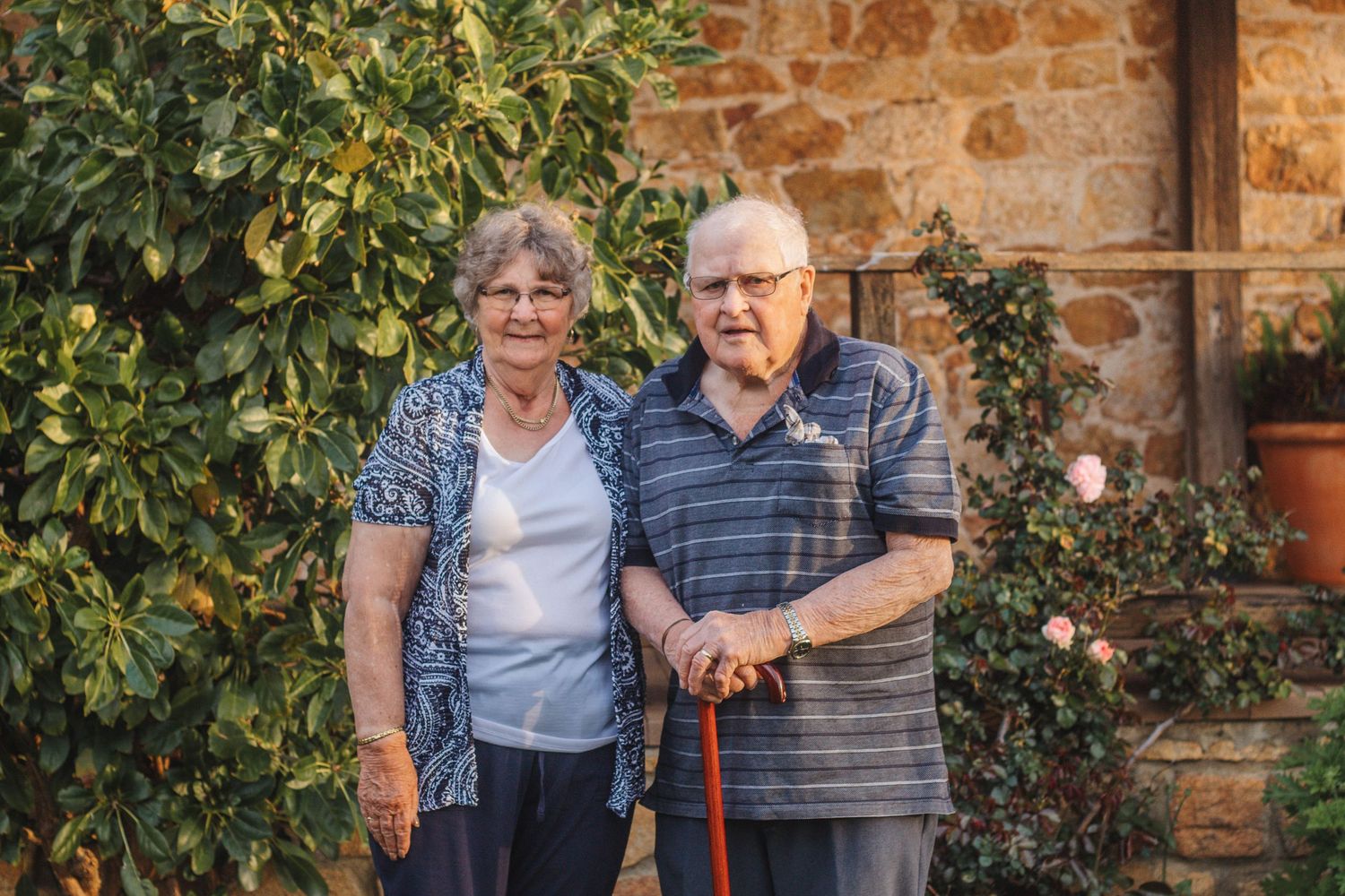 Elderly couple standing together against rustic stone wall with greenery.