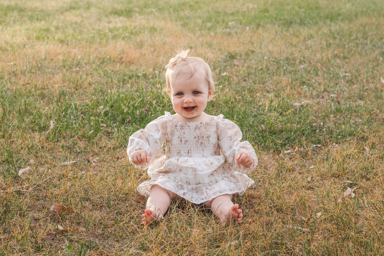 Small baby in a floral dress sitting on grass with a happy expression.
