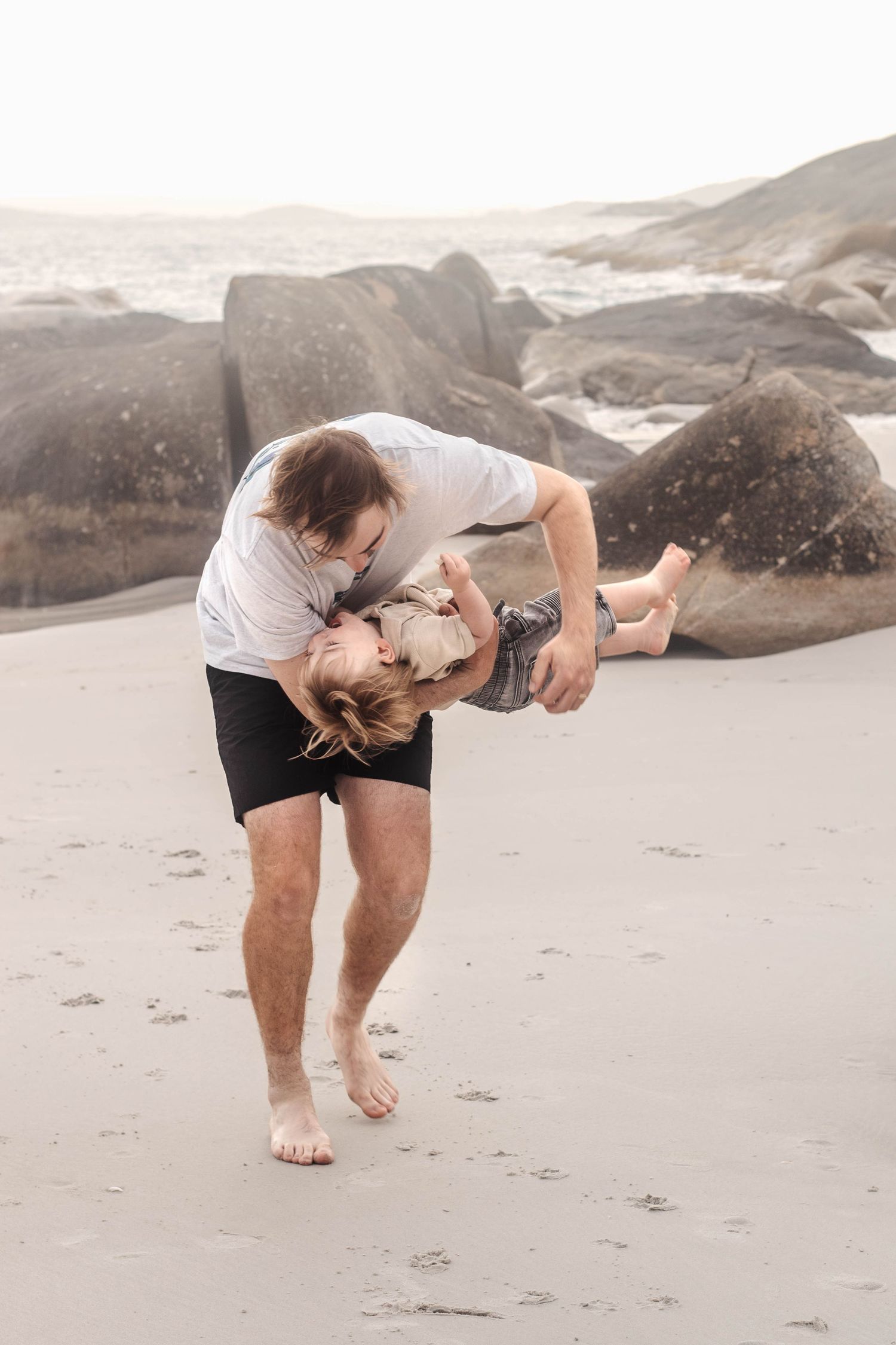 Playful interaction between adult and child on a beach with large rocks in the background.