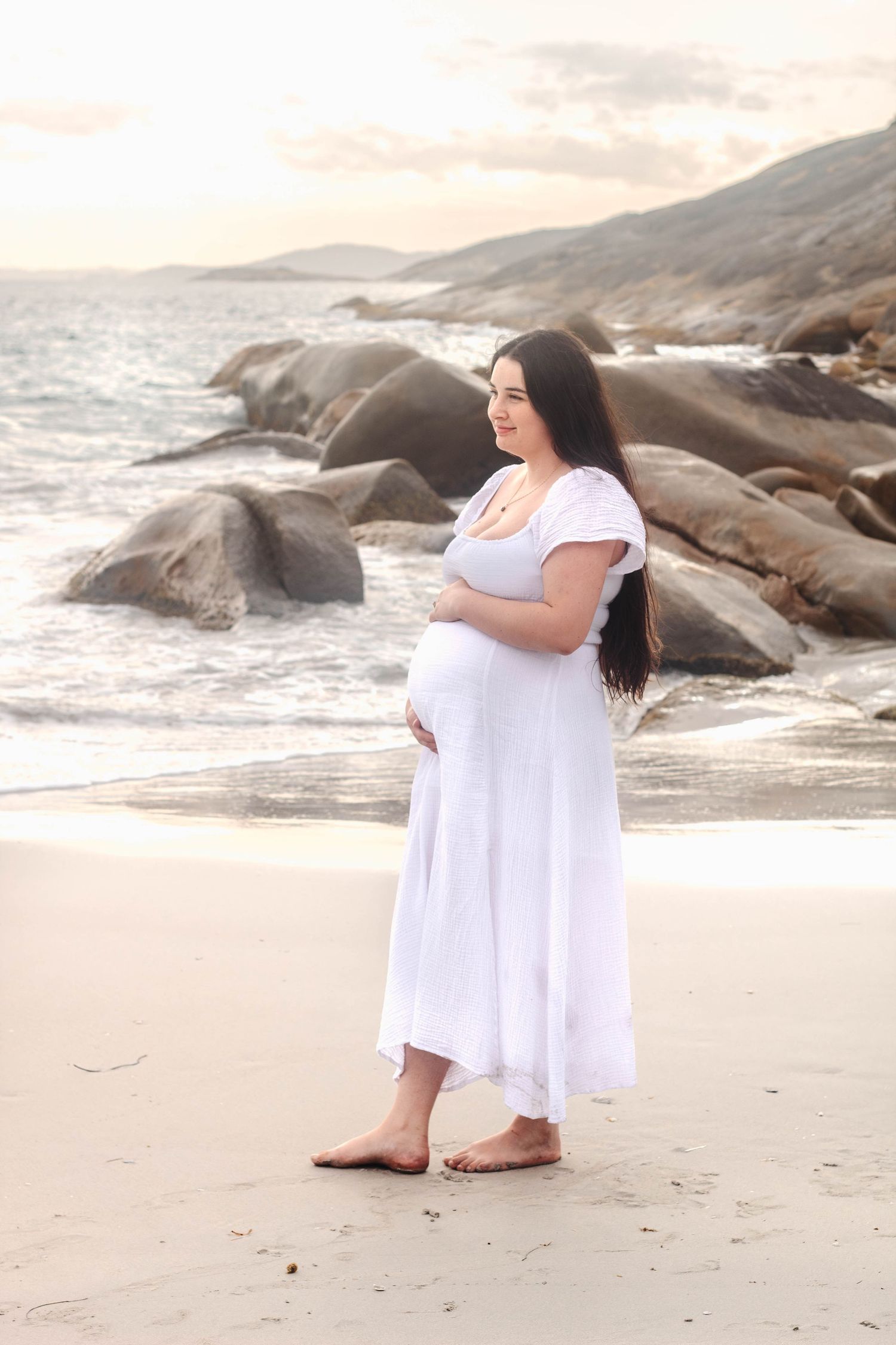 Gentle maternity portraits captured on a peaceful beach with rugged coastal rocks.