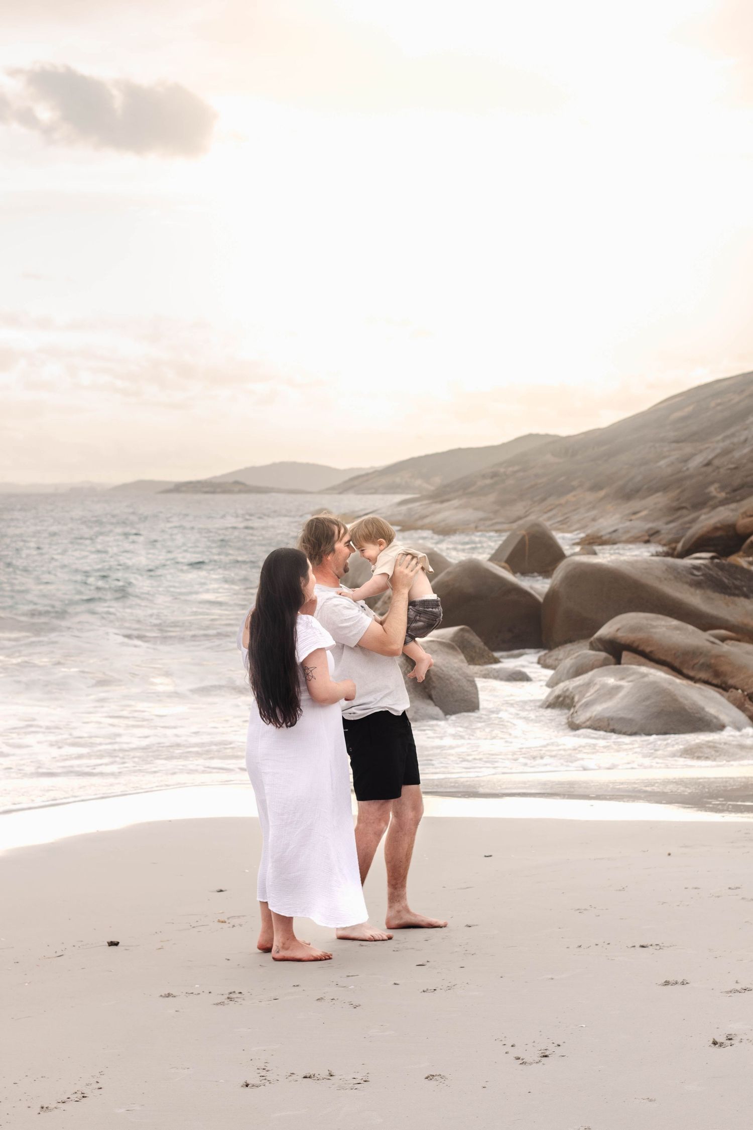 A couple performs a romantic dance lift on a sandy beach with coastal rocks and mountains.