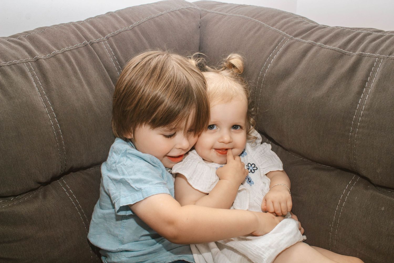 Two children snuggling and smiling together on a brown leather couch.