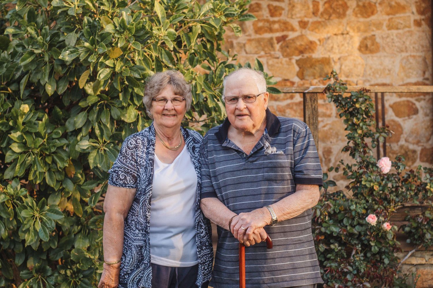Senior couple posing together outdoors against a stone wall backdrop with lush greenery.