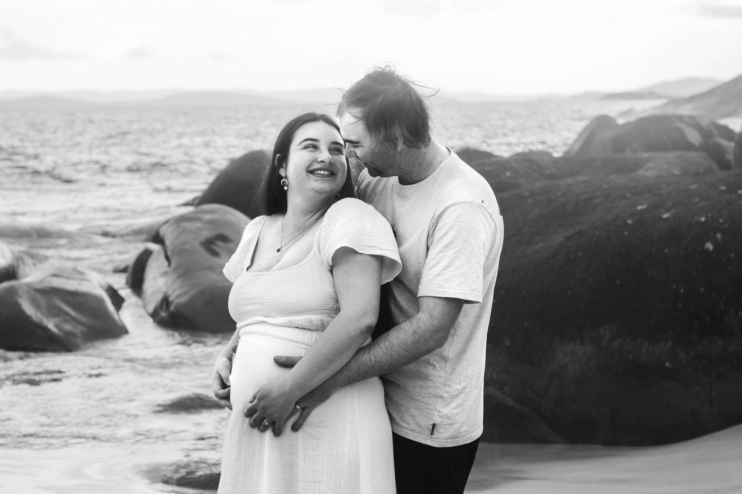 Black and white photo of expecting parents sharing intimate moment on beach.