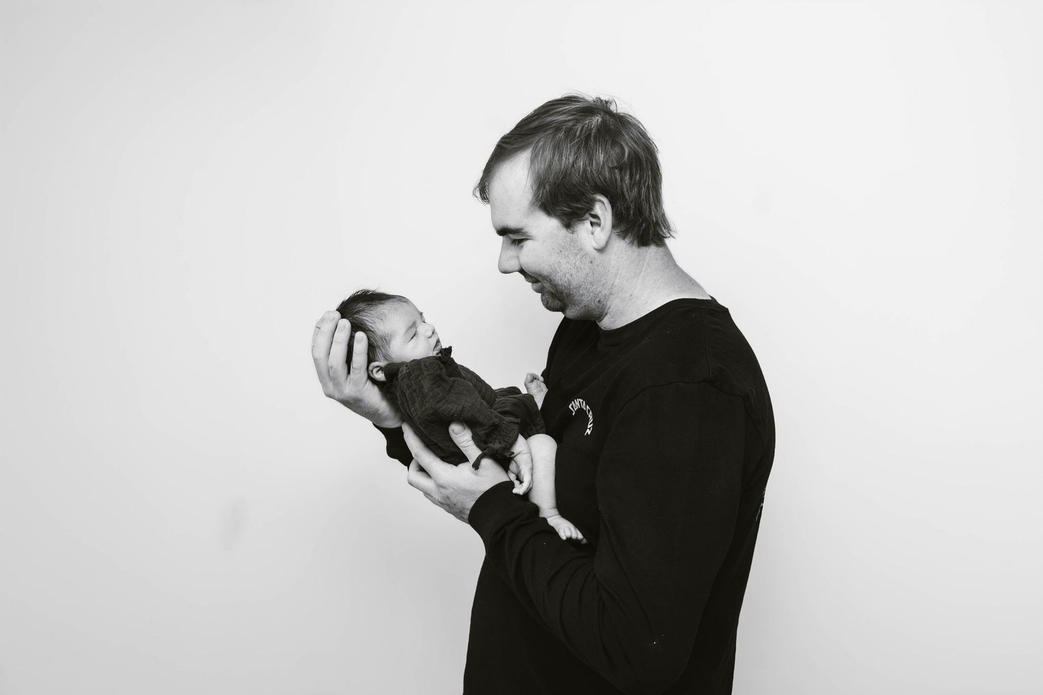Person in black sweater holding tiny newborn against white background in black and white photo.