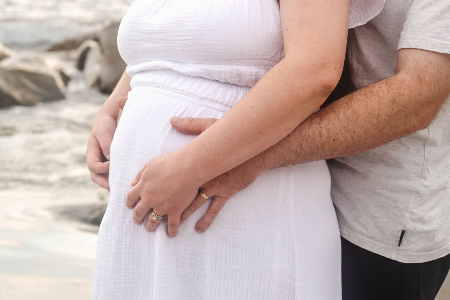 Close up of hands cradling pregnant belly in white dress on beach.