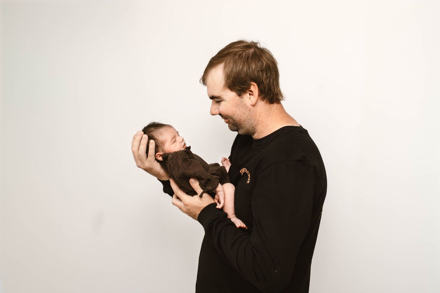 Person in dark sweater holding small newborn against white studio background.