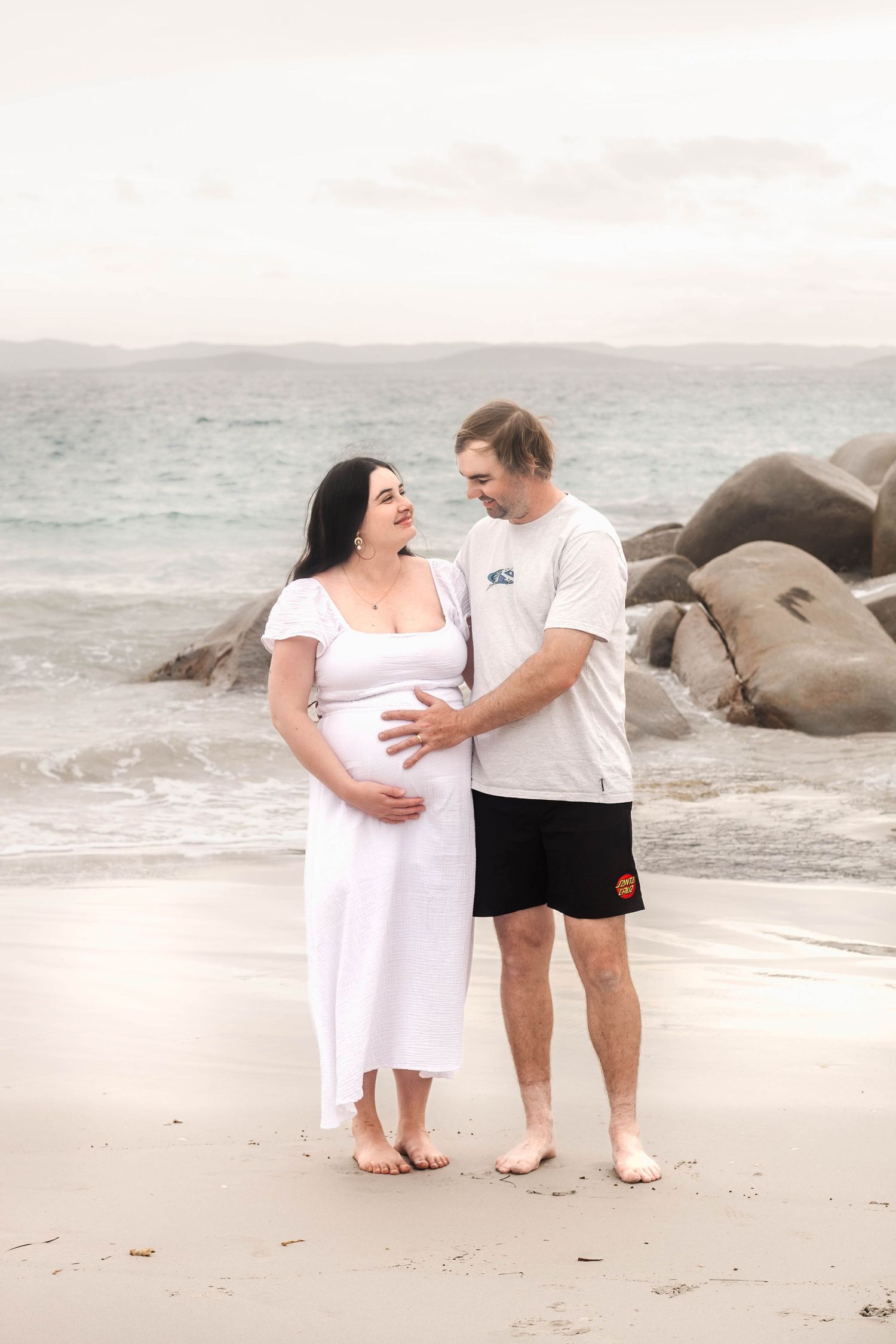 Couple standing together on sandy beach with rocky coastline in background.