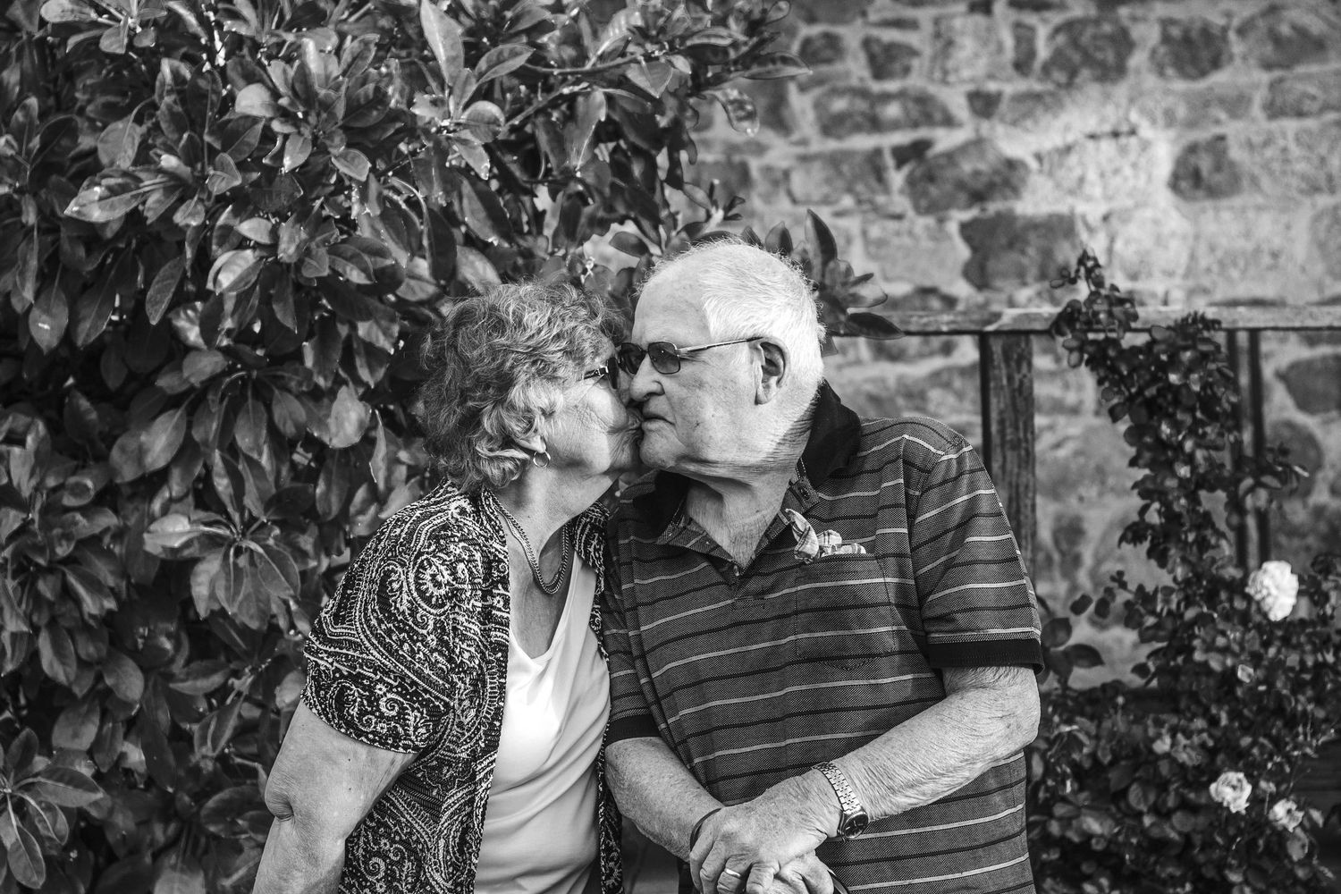 Black and white capture of an intimate moment between elderly couple by a garden wall.