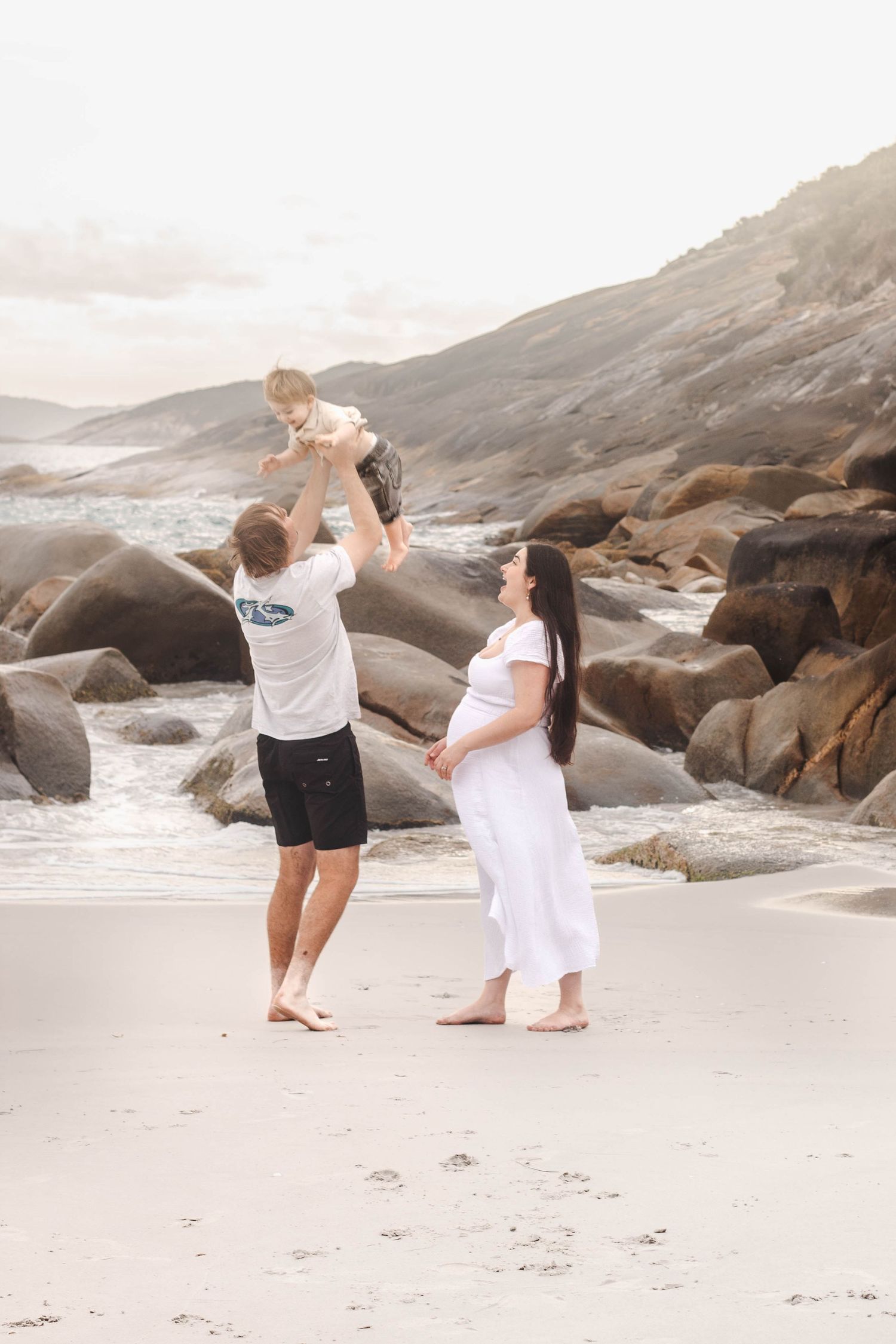 Parents playing with their child on a sandy beach with dramatic rocky coastline.