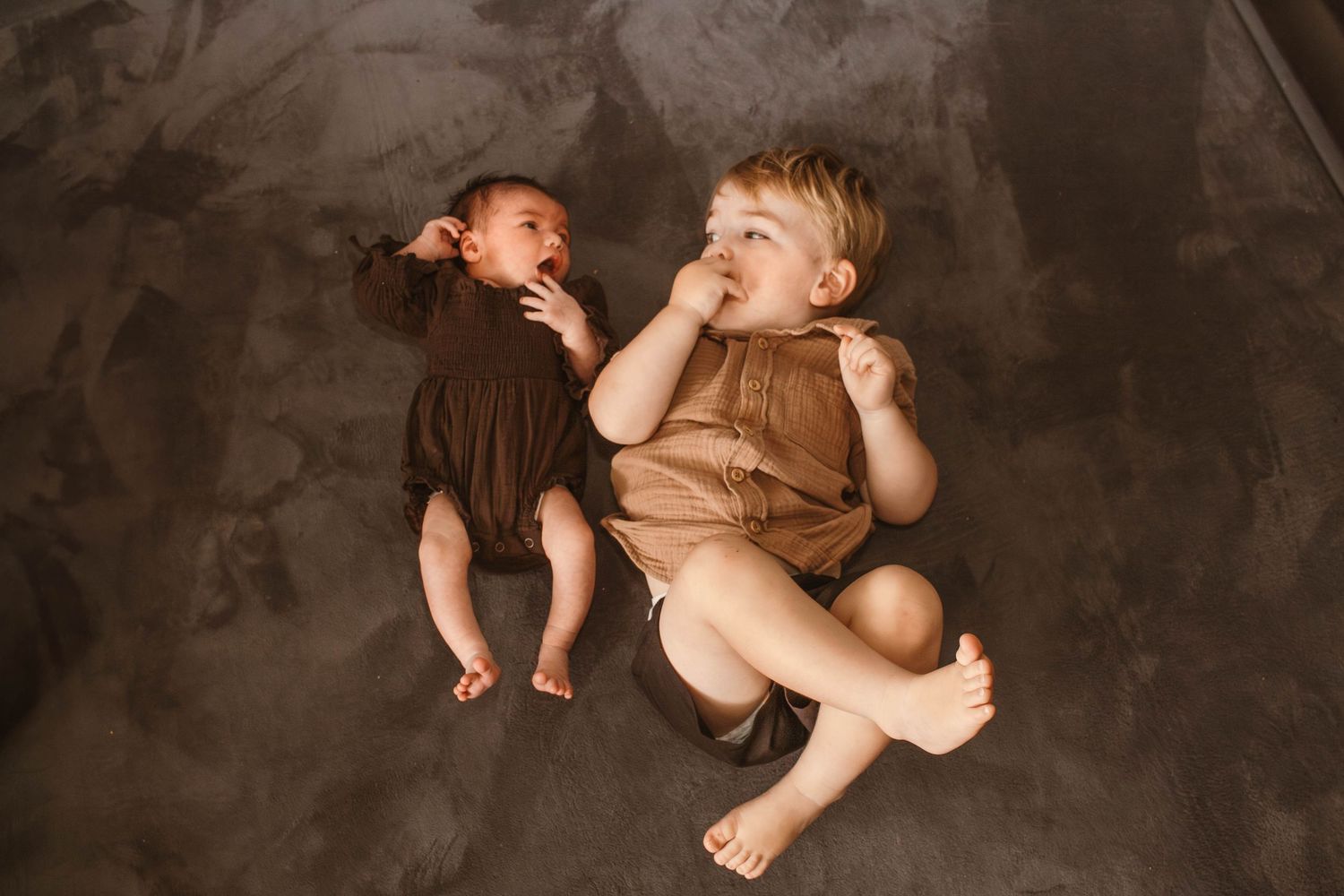 Two babies lying on dark backdrop wearing contrasting brown and beige outfits laughing together.
