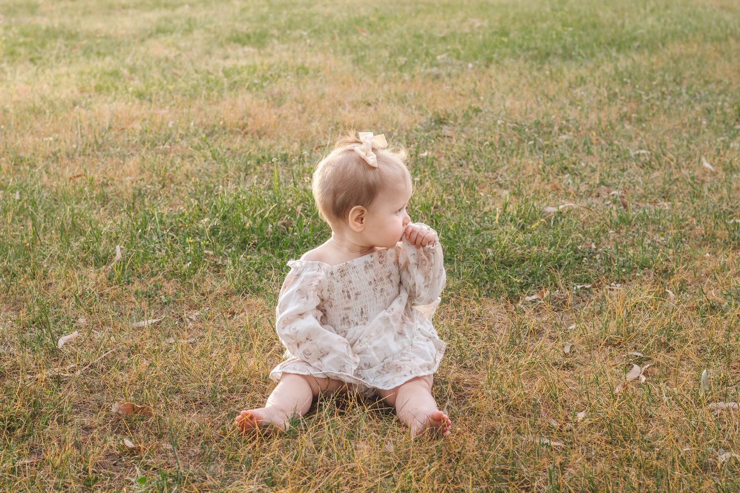Baby in vintage-style dress sitting on dry grass with curious expression.