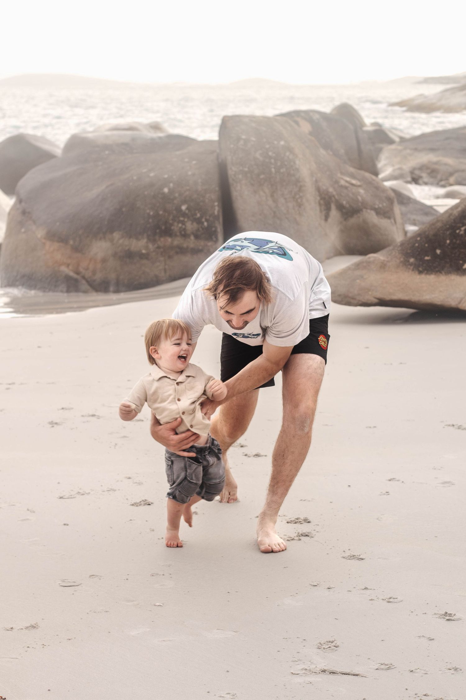 Joyful beach playtime with adult helping small child walk on sand near large boulders.