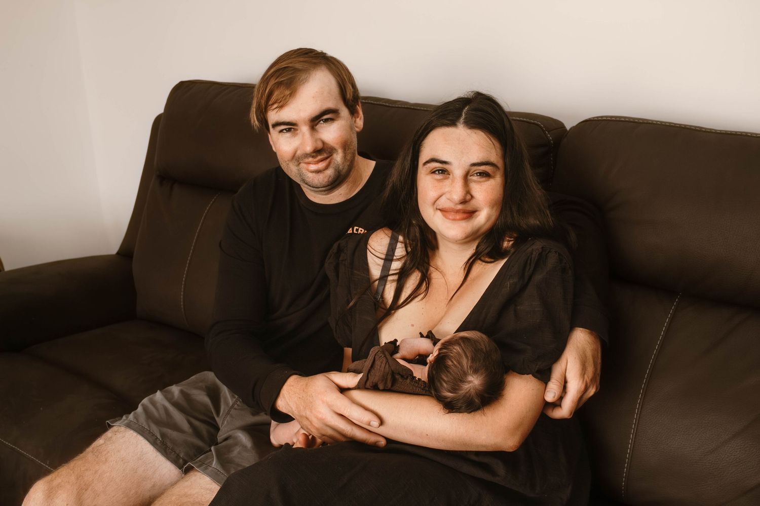 A couple sits on a brown couch while holding their newborn baby in a warm, intimate setting.