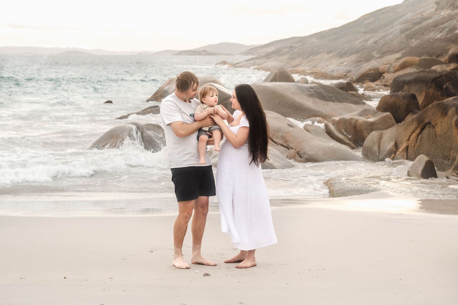 A couple shares a tender moment with their baby on a rocky beach at sunset.
