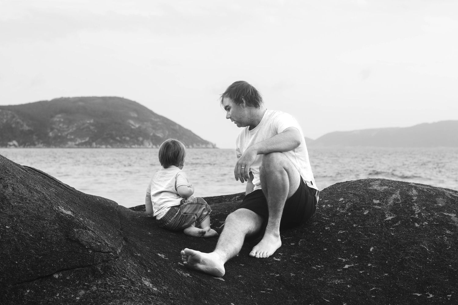 Parent and child sitting together on rocky ocean overlook in black and white.