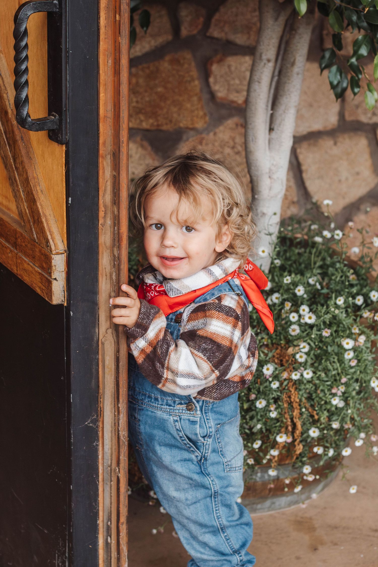 Toddler in plaid shirt and jeans stands playfully by wooden door with stone wall background.
