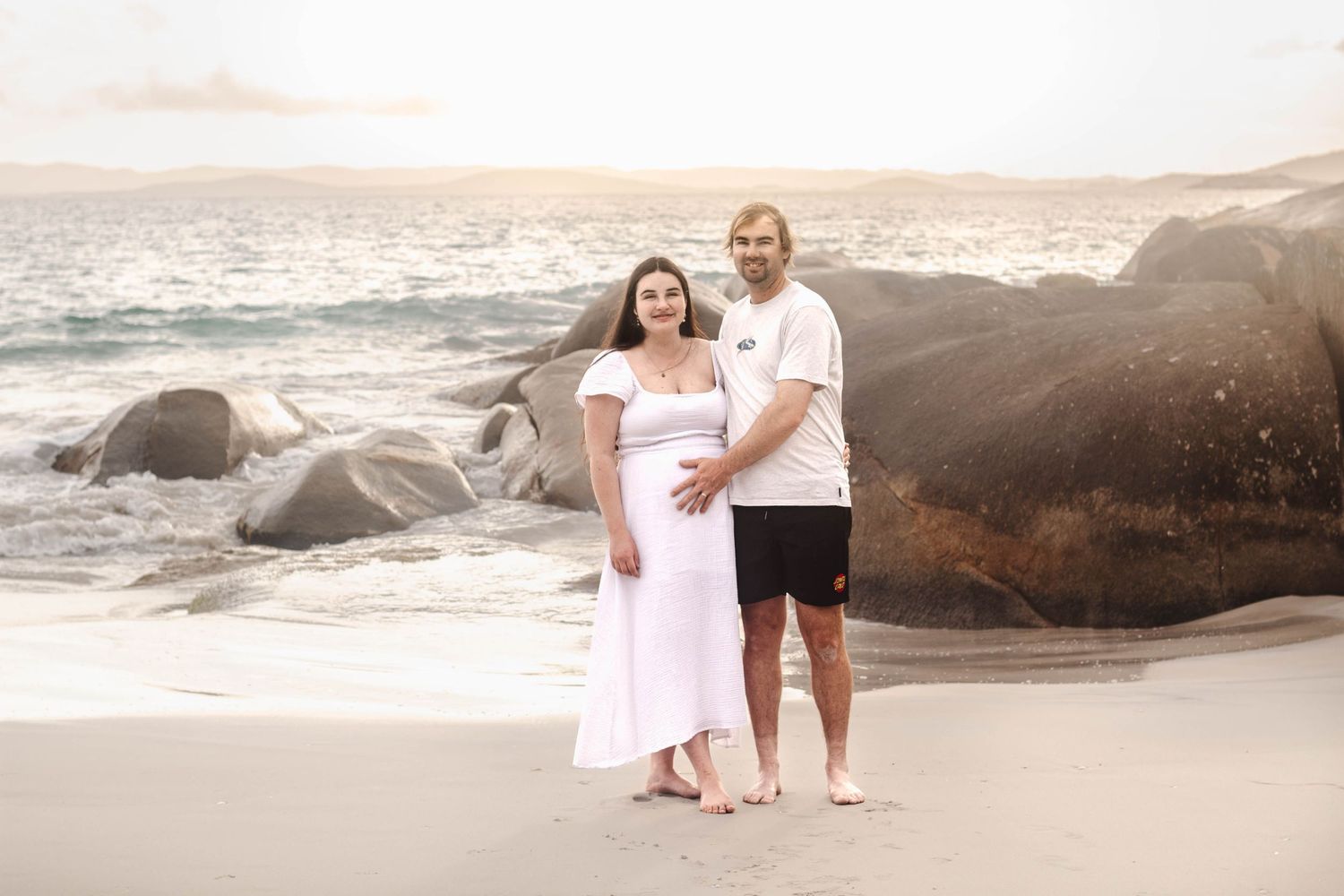 Expectant couple stands together on beach with dramatic rocky coastline during sunset.