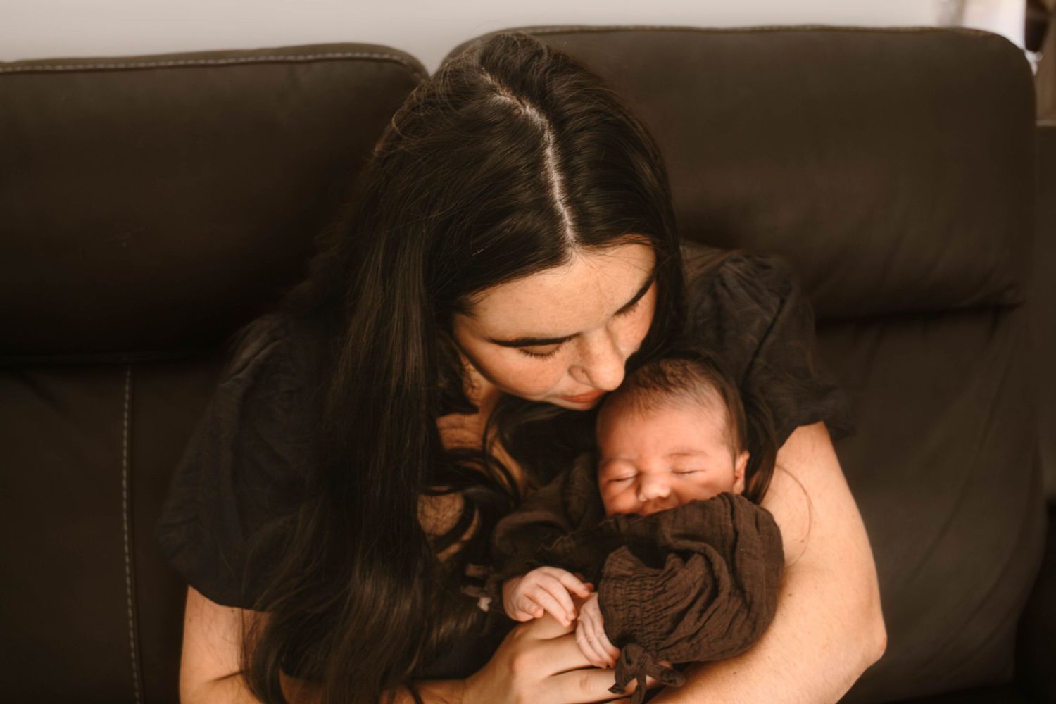 Mother wearing dark clothing cradles sleeping newborn on brown leather sofa.