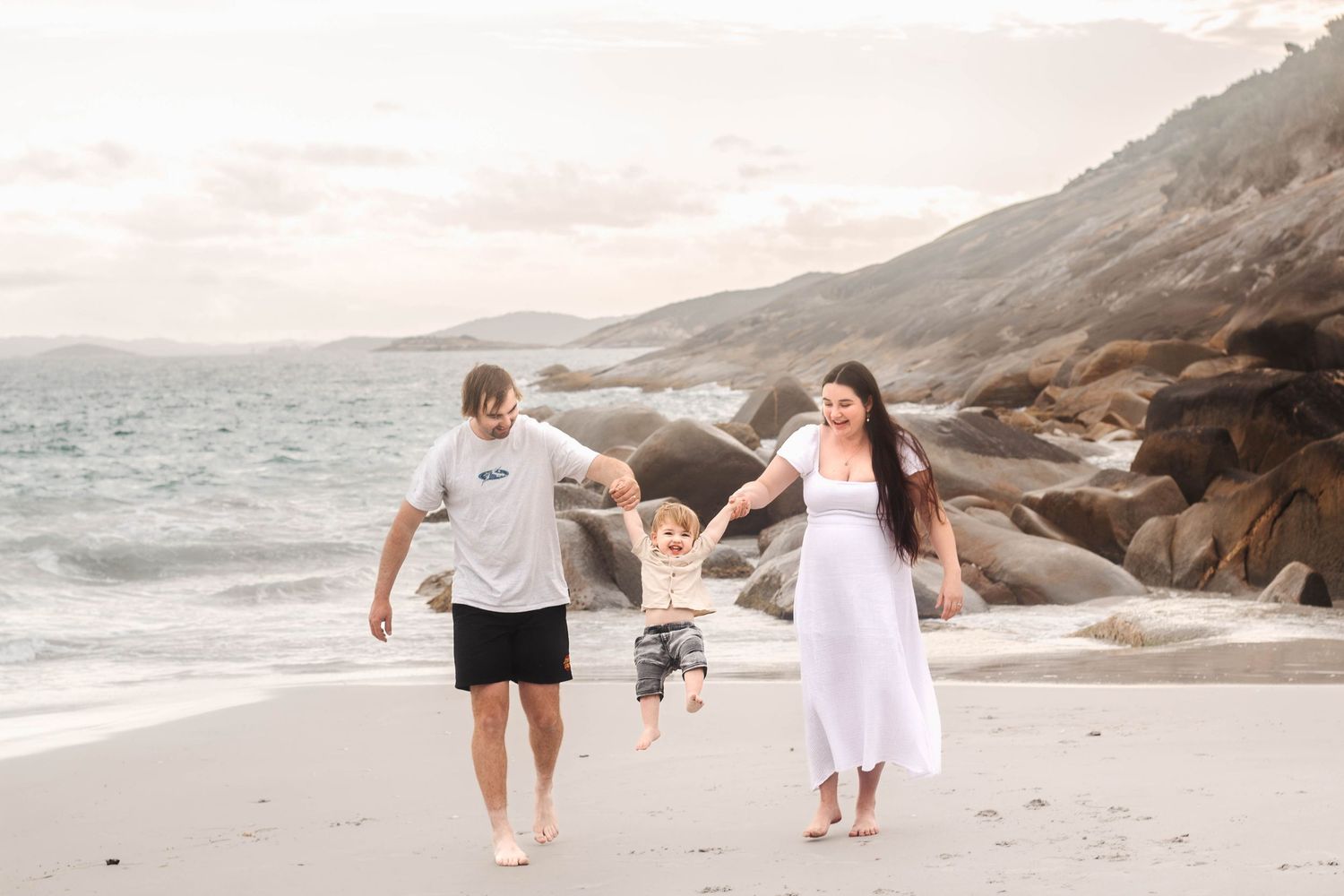 Family holding hands while walking along a scenic beach with rocky cliffs.