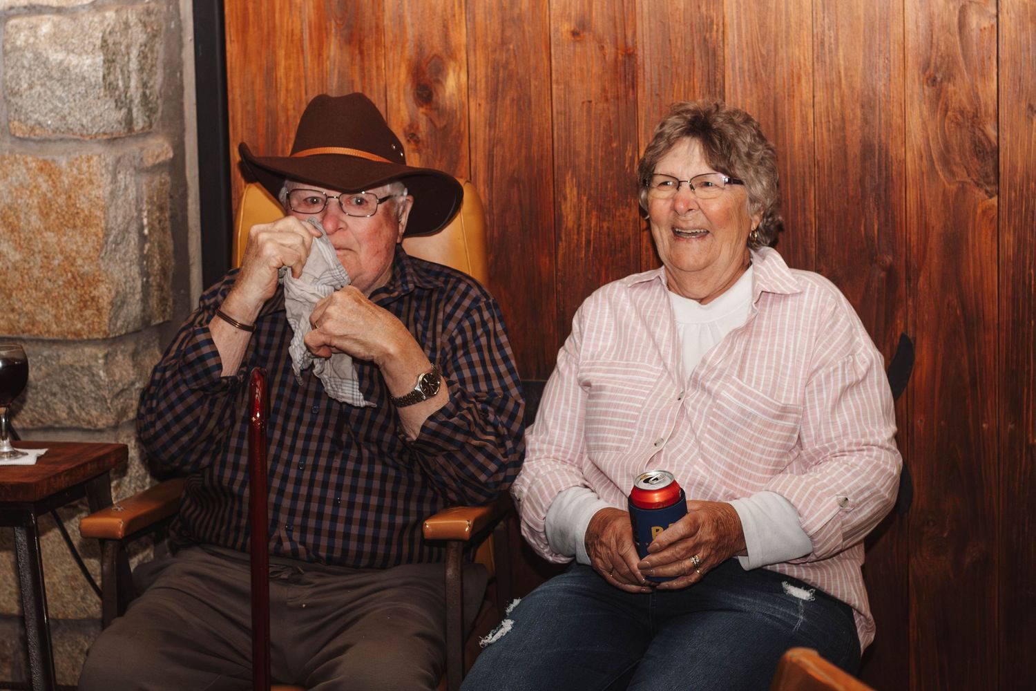 Two friends enjoying beverages while sitting together against a stone and wooden wall backdrop.