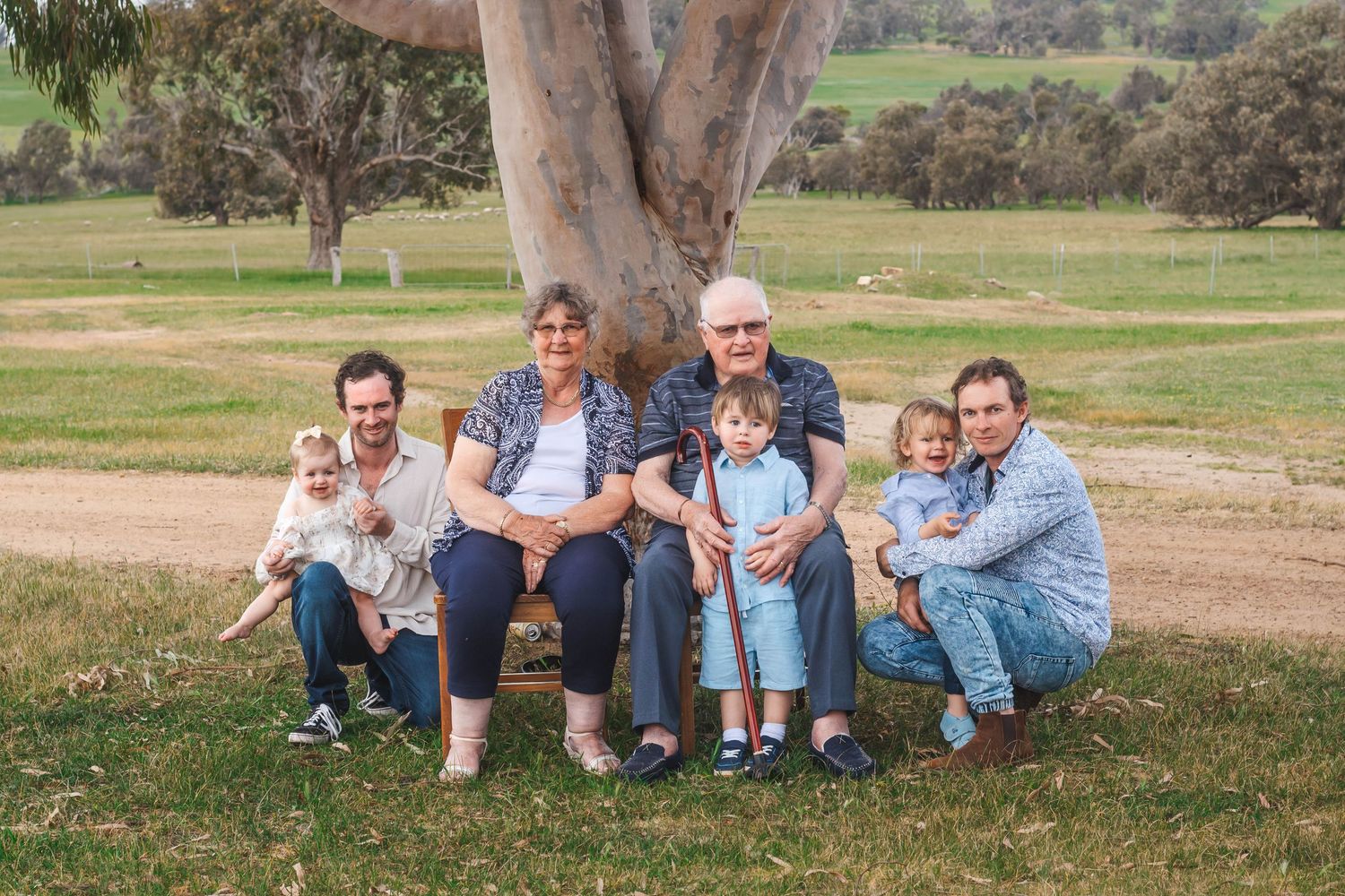 Extended family portrait sitting together under large gum tree in countryside.