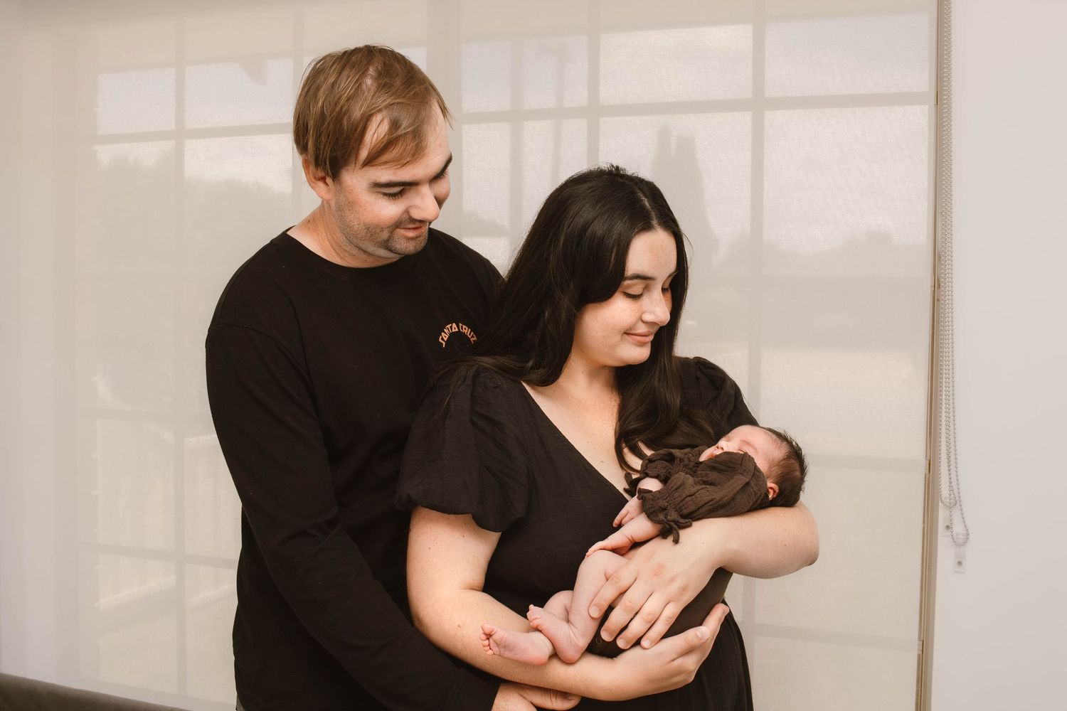 A couple dressed in black shares a tender moment with their newborn by a bright window.