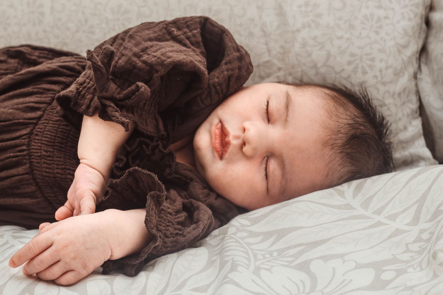 Sleeping baby in brown quilted dress lying on light colored bedding.