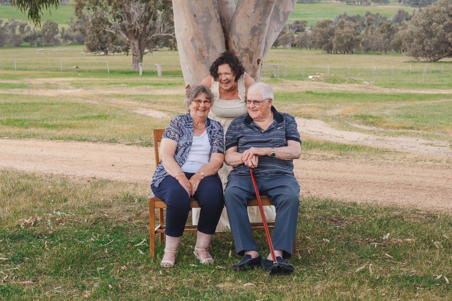 Couple relaxing on wooden chairs under a large gum tree in Australian countryside.