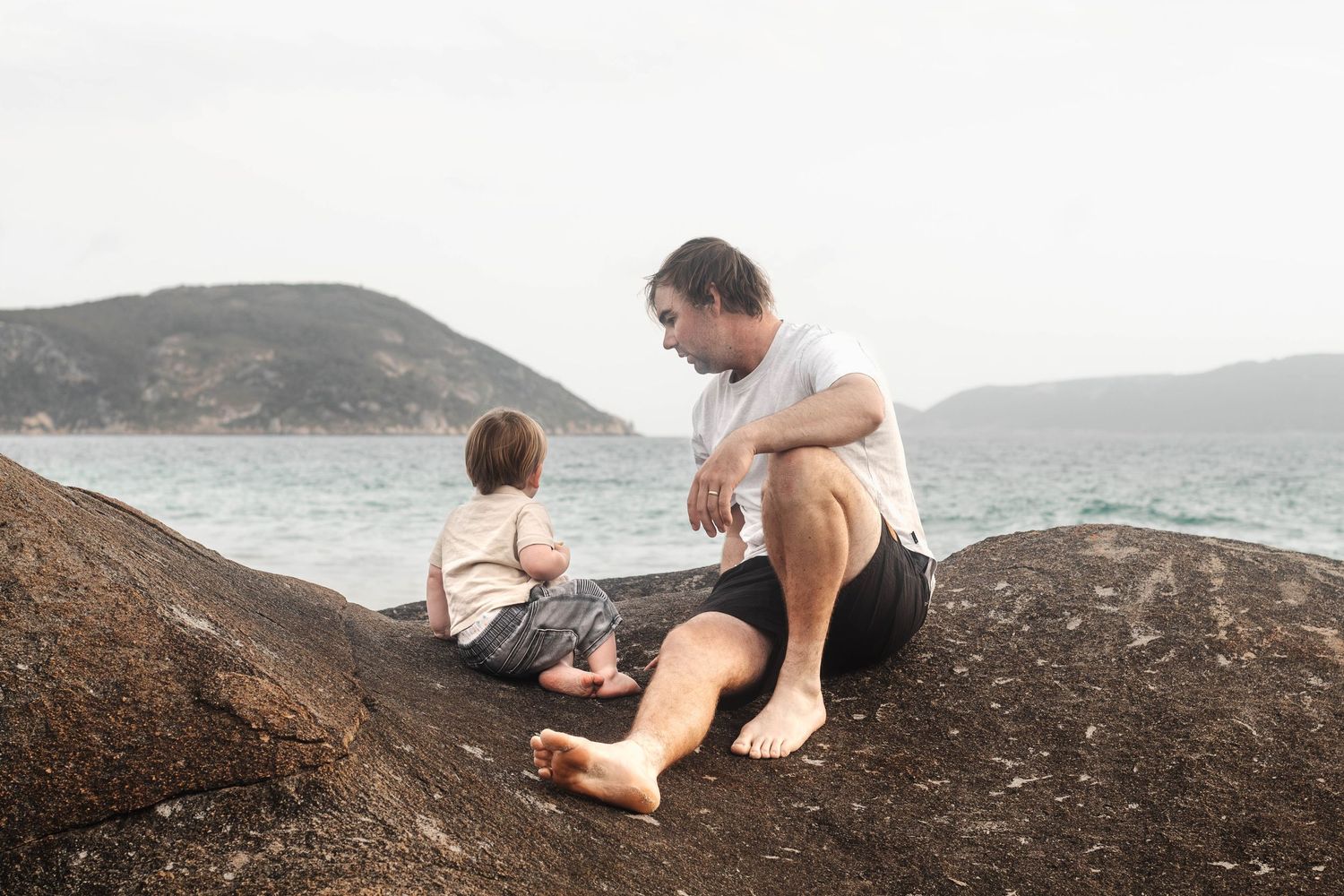 Father and child spend quality time sitting on coastal rocks overlooking ocean.