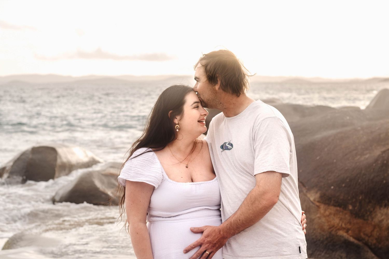 Expecting couple shares a loving moment on a beach with rocky coastline at sunset.