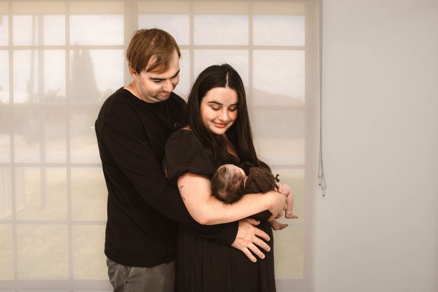 A couple dressed in black holds their newborn baby in front of a window with natural light.