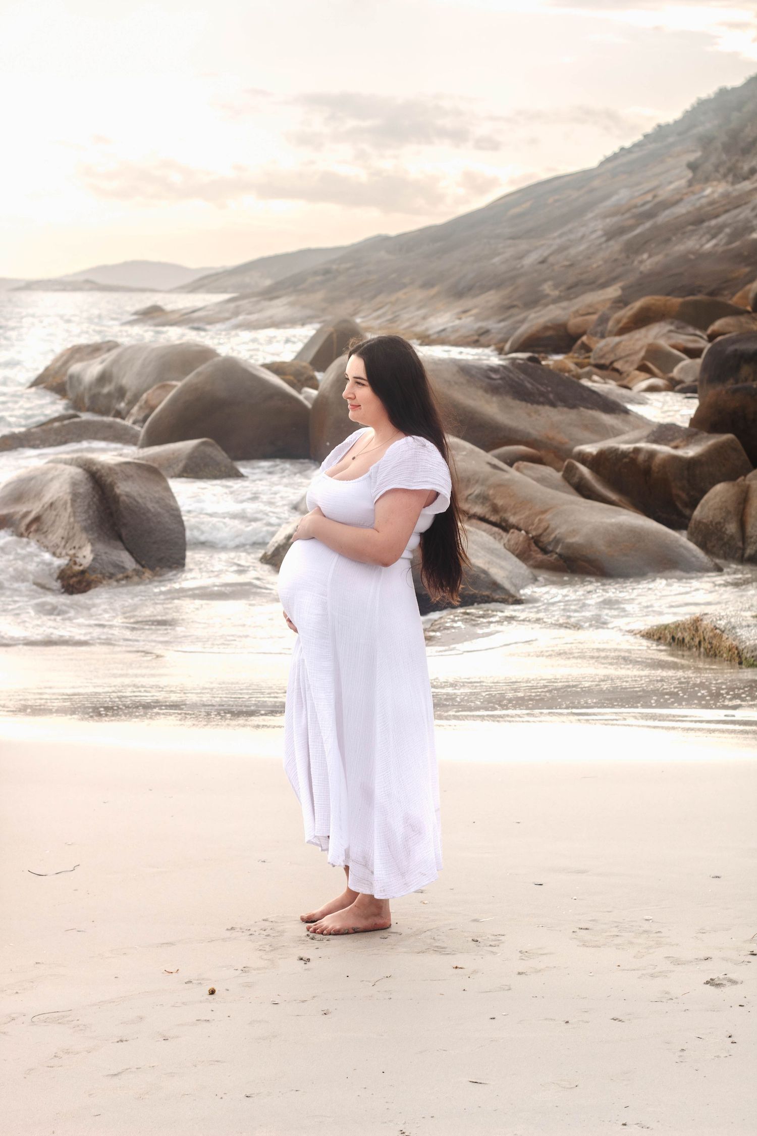Serene maternity photoshoot on a sandy beach with dramatic rocky coastline at dusk.
