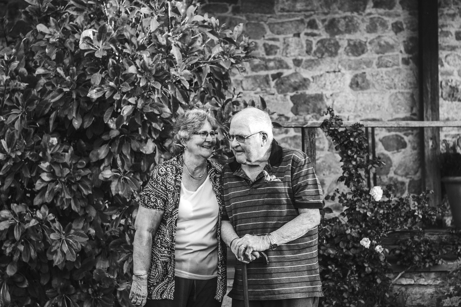 Black and white photo of a senior couple standing together by a stone wall with climbing plants.