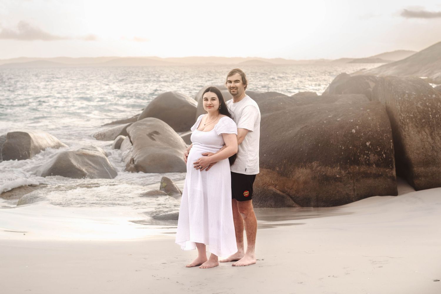 Couple embraces on a rocky beach at sunset during maternity photoshoot.