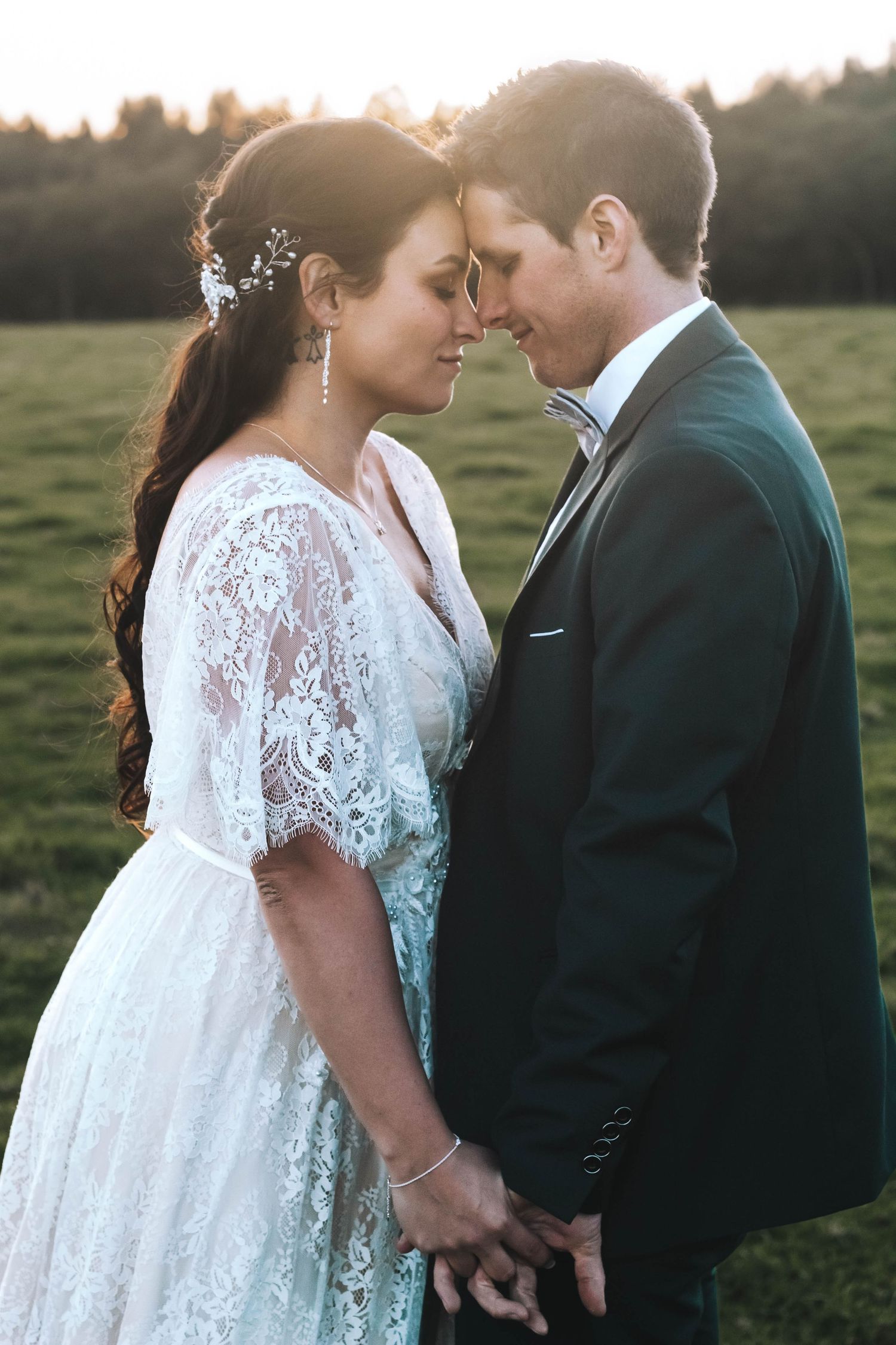 A romantic wedding portrait of a couple in a field at sunset, the bride wearing a lace dress and delicate hair accessory.