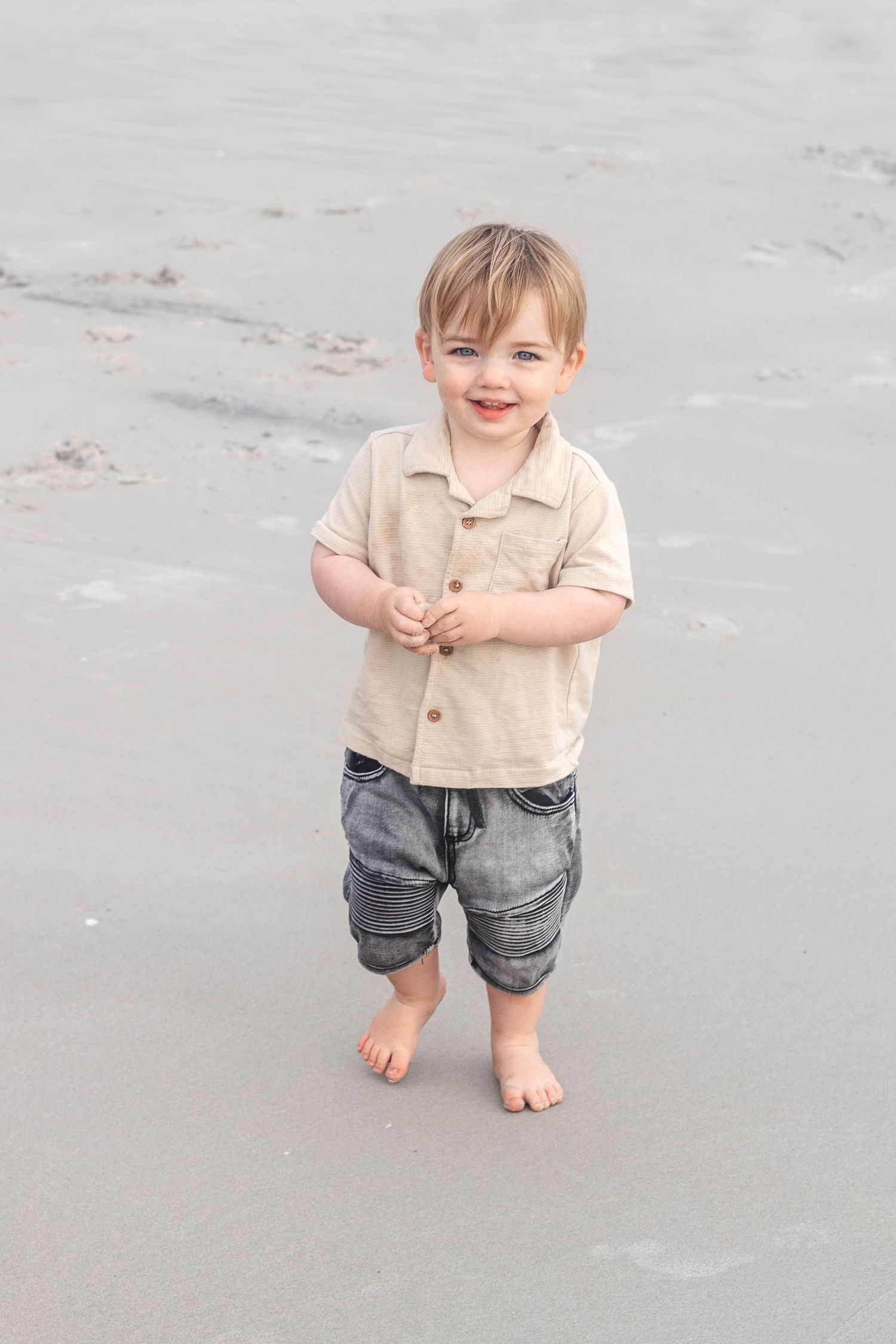 Candid toddler portrait on sandy beach wearing casual beige shirt and denim shorts.