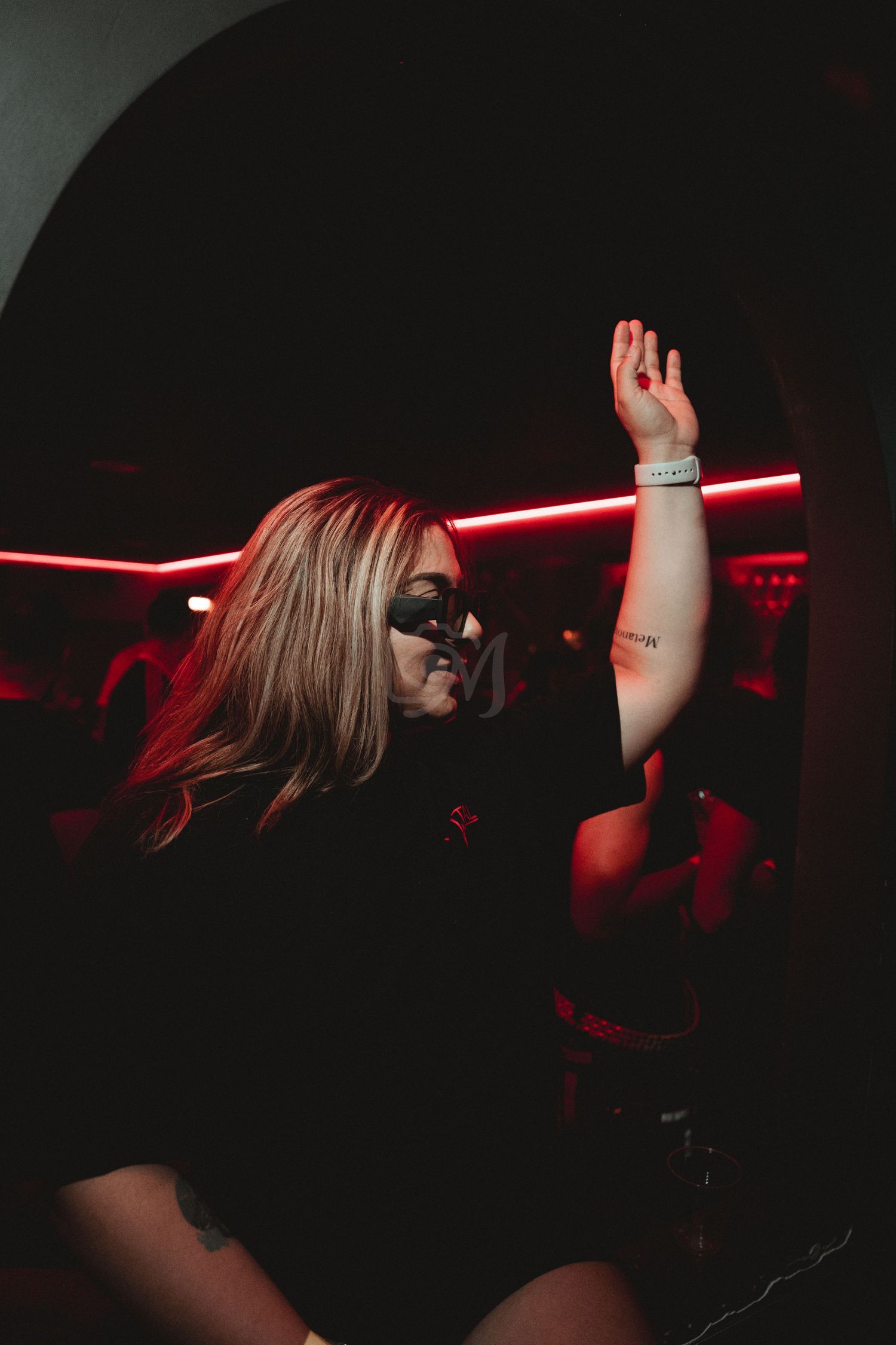 Dancer in sunglasses raises arm while performing under red club lights.
