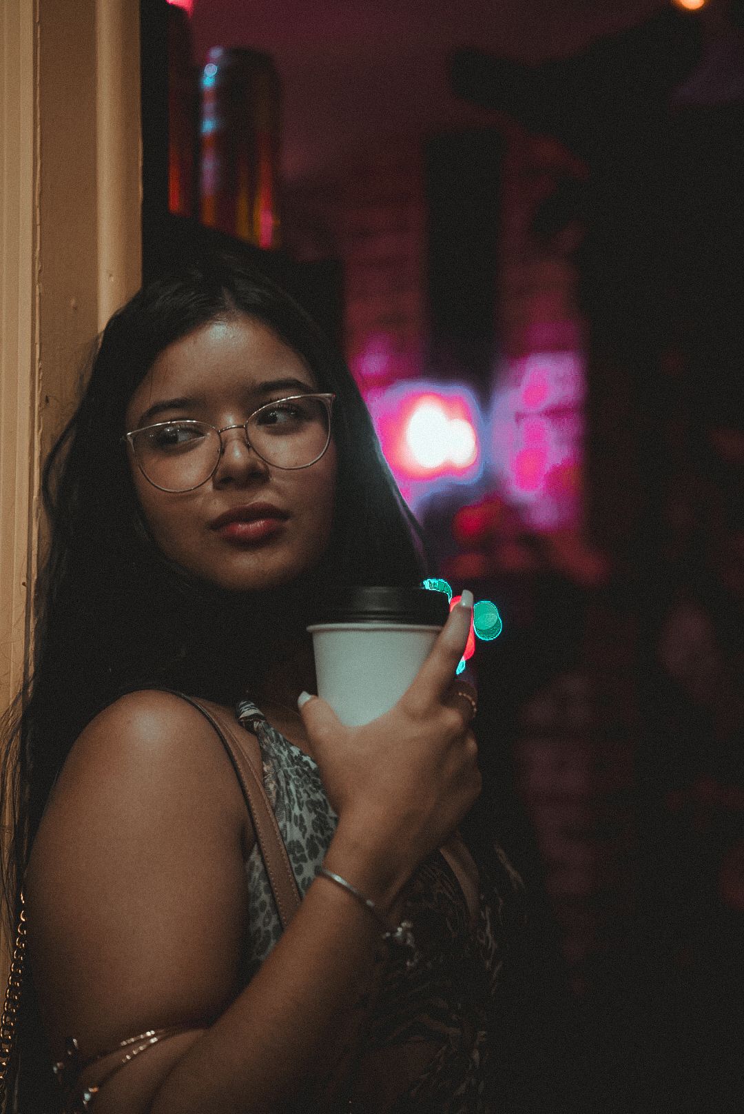 A young woman in glasses holds a coffee cup against a dark background with pink neon lights.