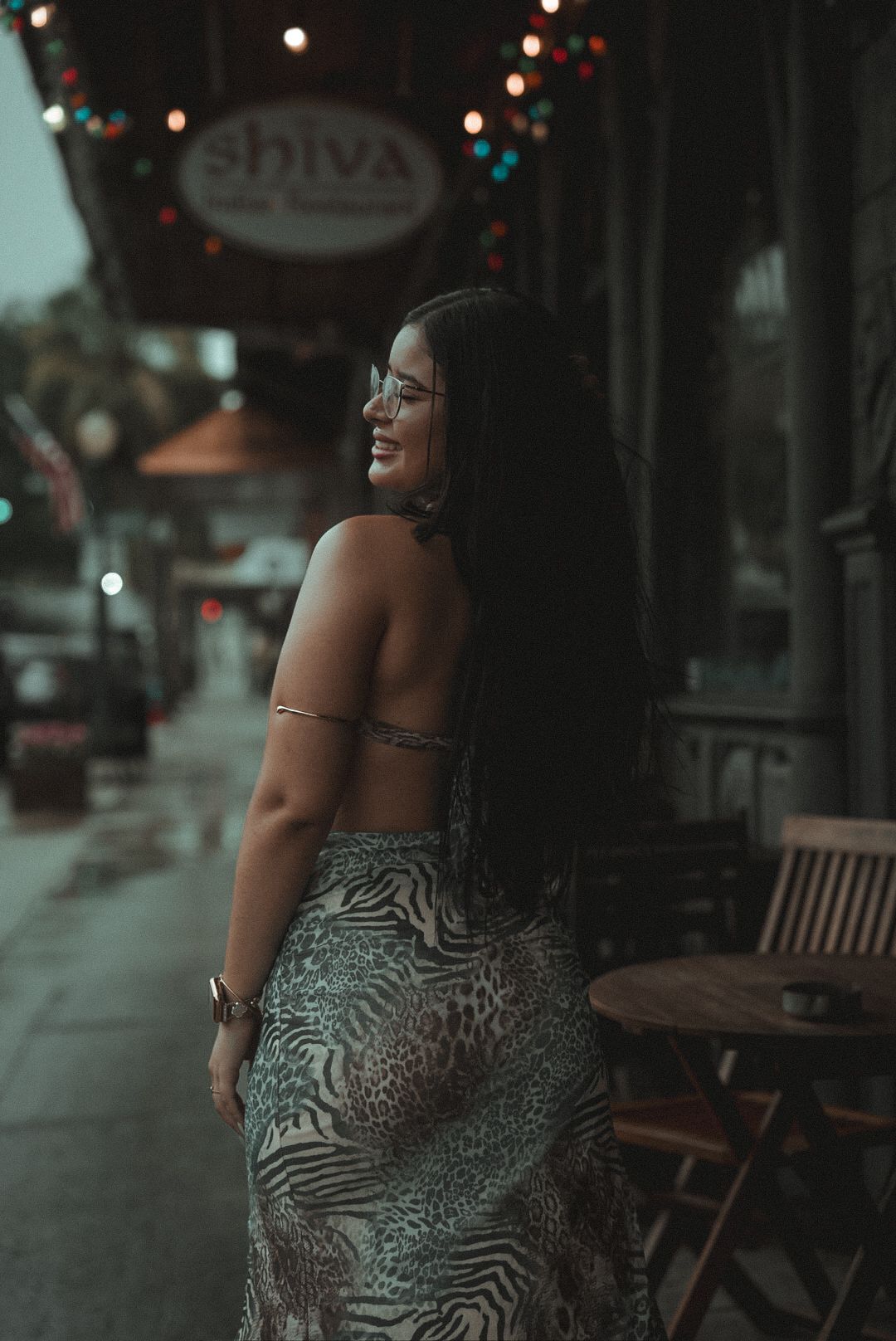 Stylish person stands in evening light wearing a zebra print dress at an outdoor cafe with bokeh lights in background.