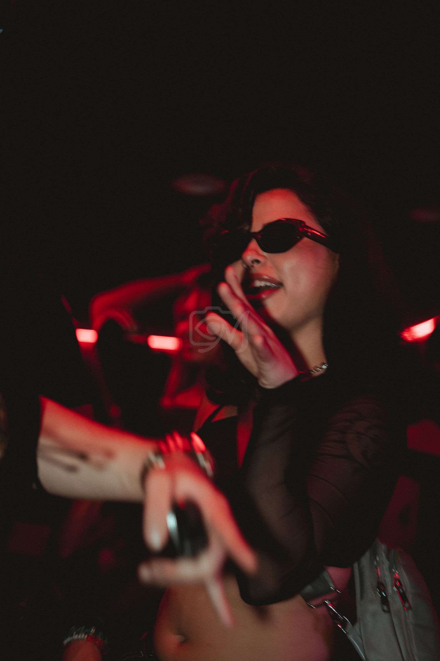 Person wearing sunglasses and black top performs in dark nightclub setting with red lighting effects.