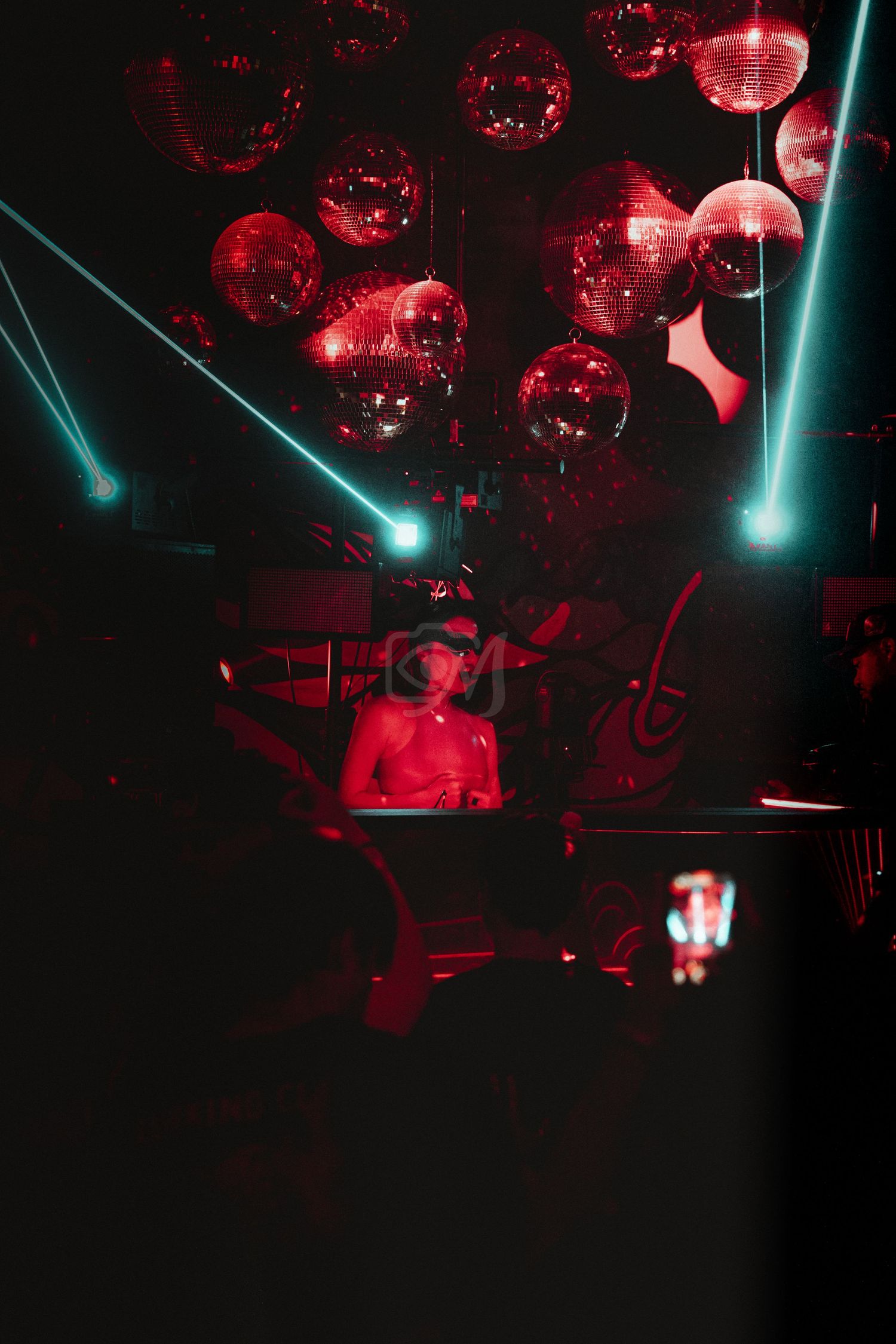 Disco balls and teal laser beams create atmosphere in a red-lit nightclub.