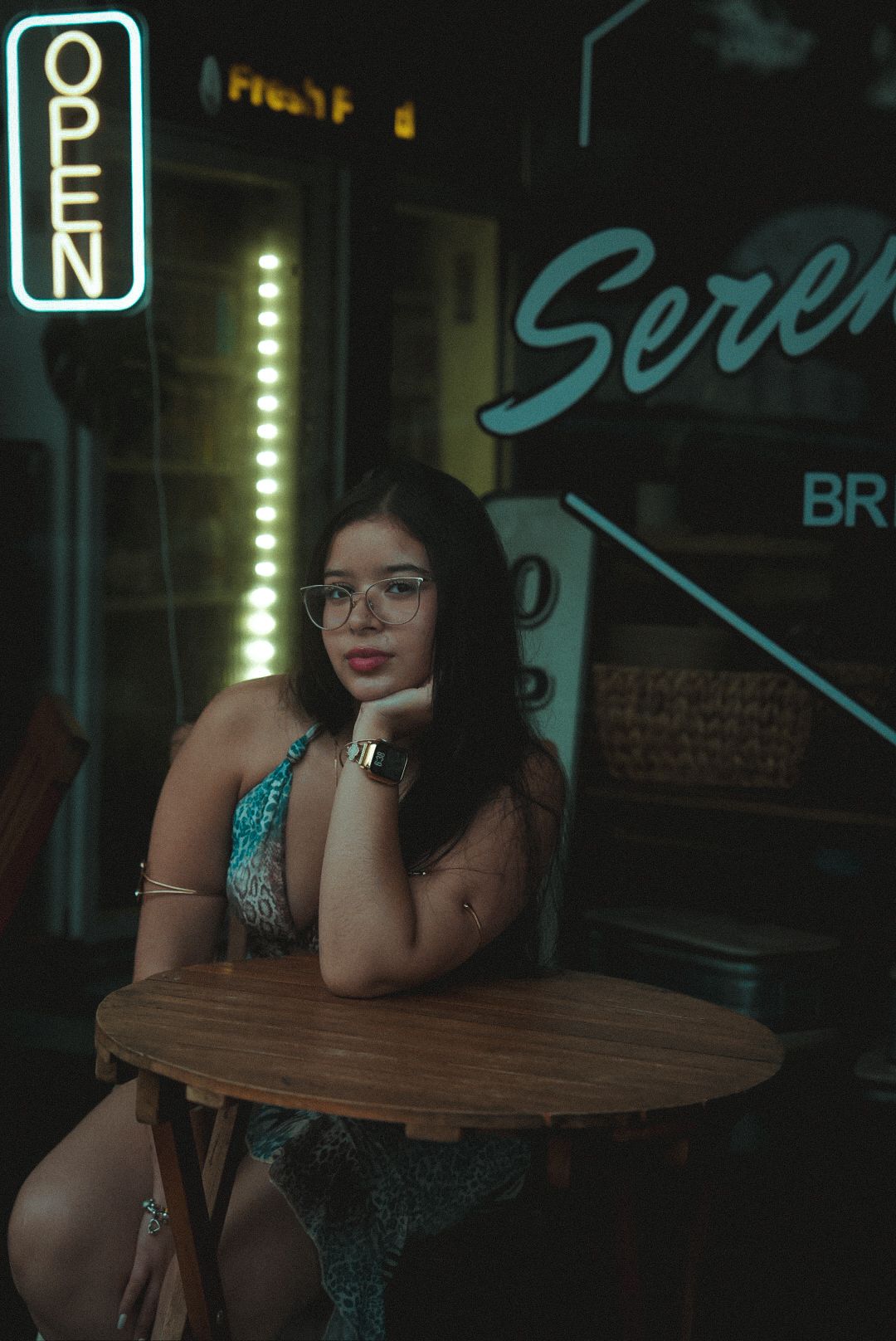 Moody nighttime portrait showcasing neon signs and a person seated at a small wooden table in a sparkly dress.