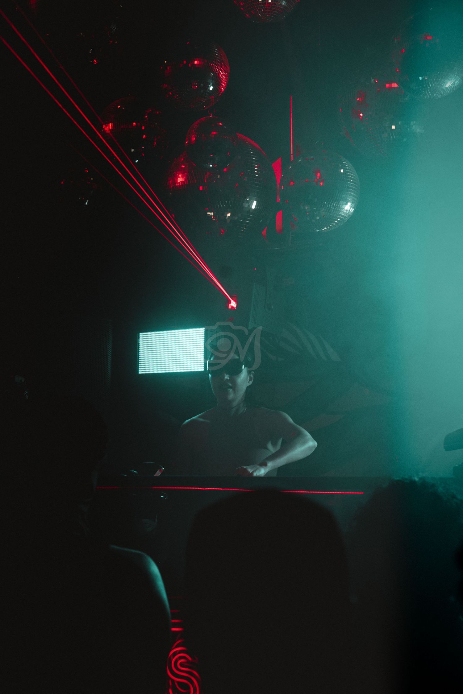 DJ performing behind decks with disco balls and red laser beams in dark atmospheric club setting.
