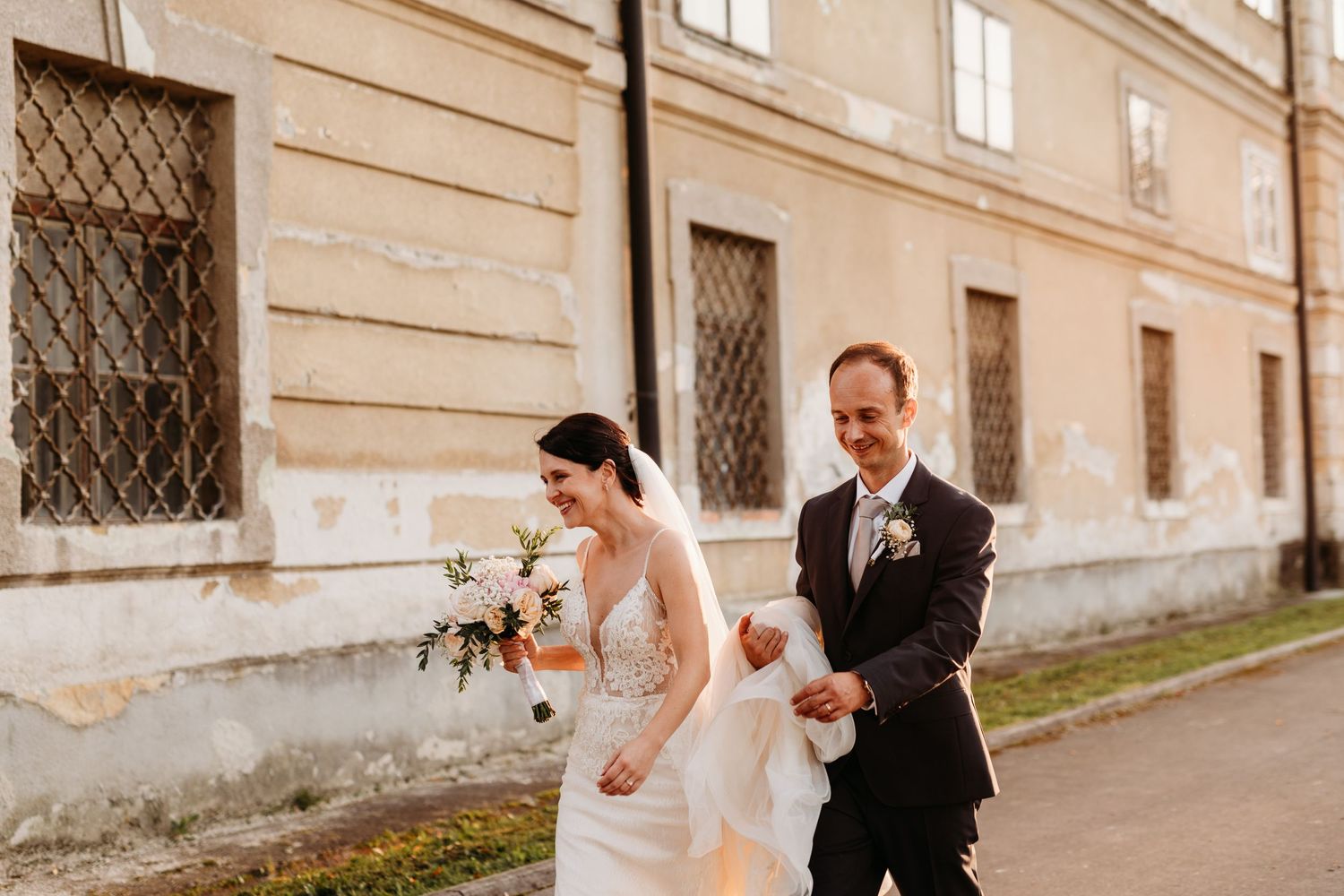A couple walking along a historic building exterior with bride holding bouquet.