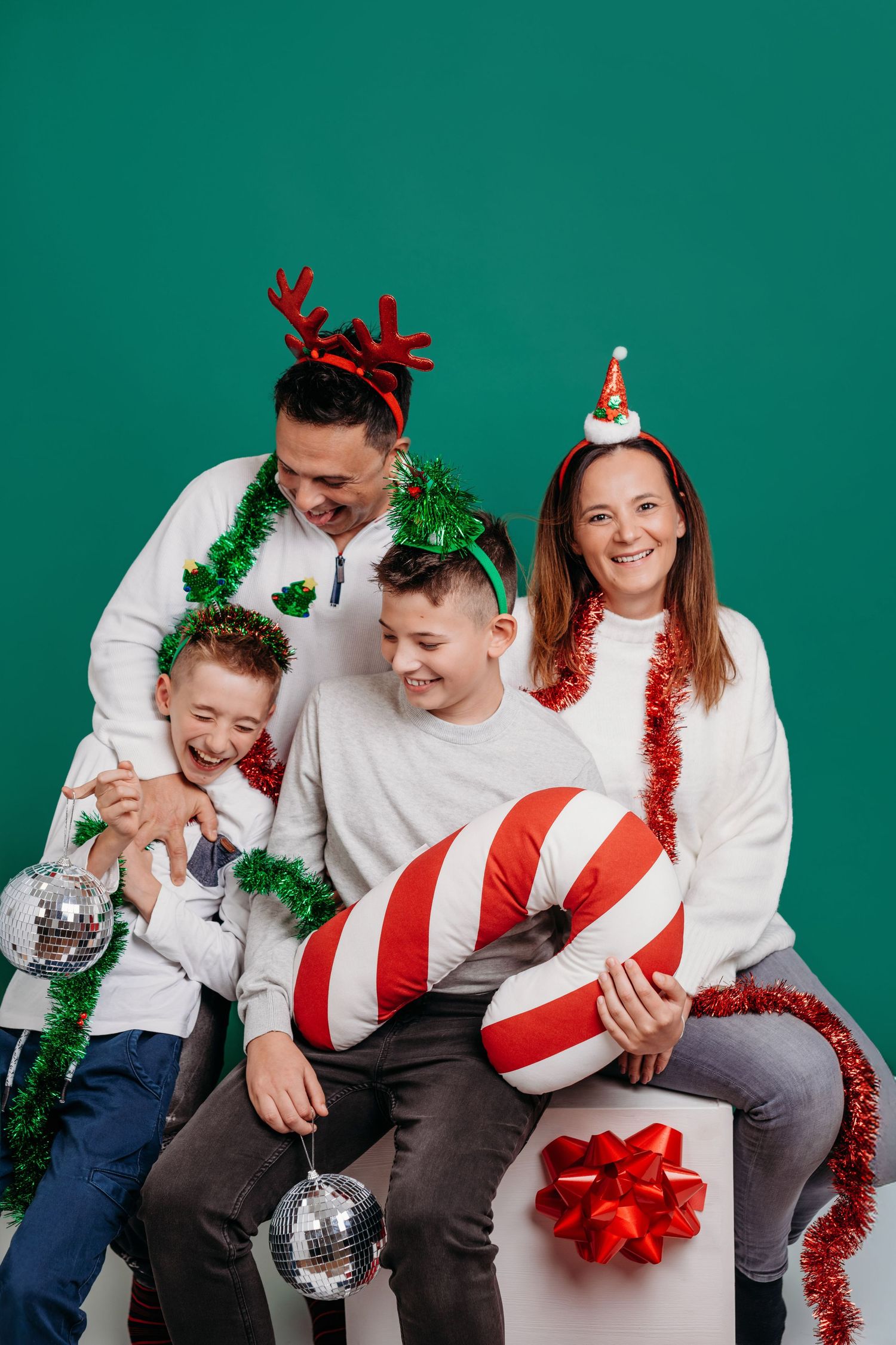 A festive family group in white shirts poses playfully with Christmas decorations and tinsel against a green backdrop.