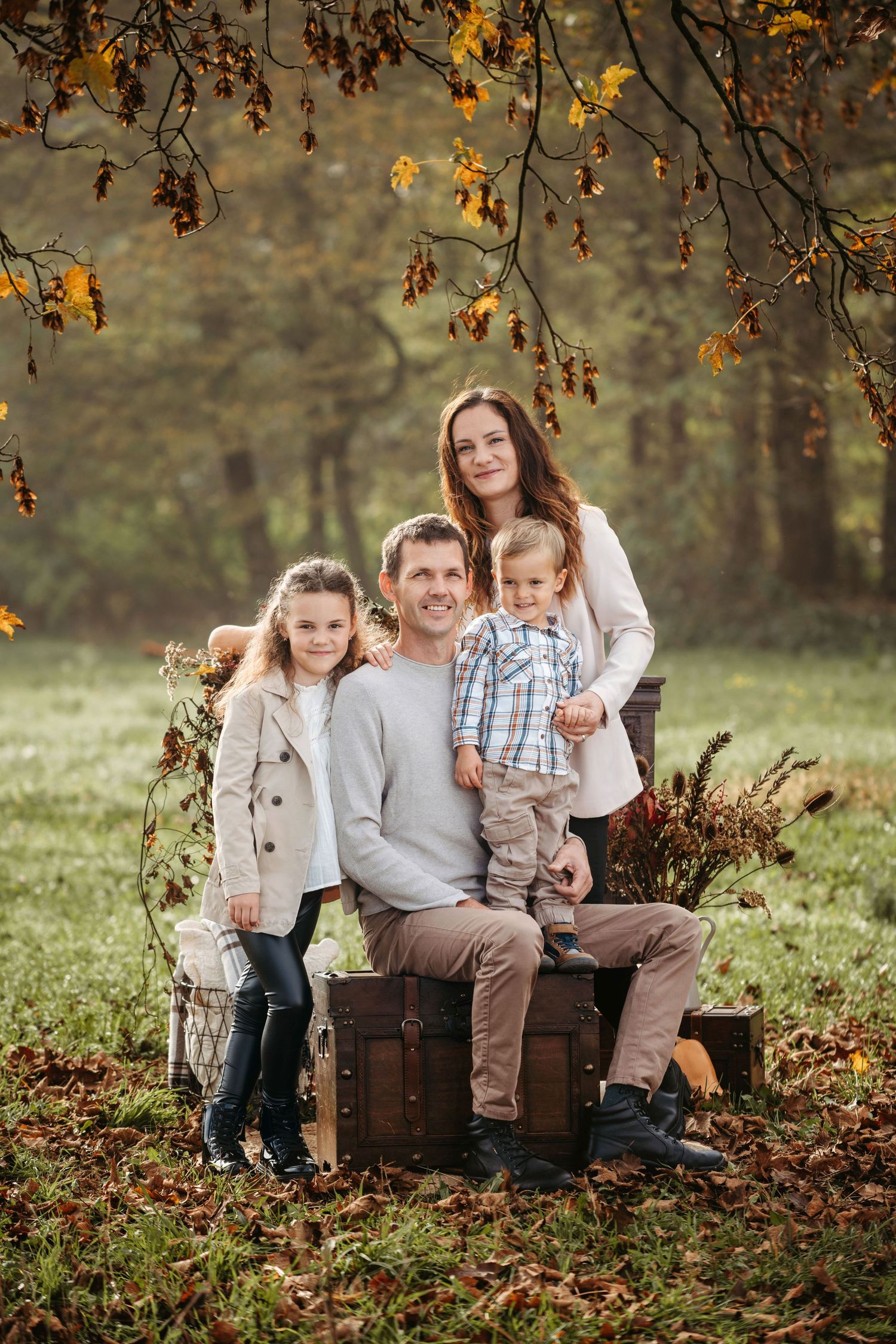 Family sits together on wooden crate surrounded by autumn leaves and golden foliage in outdoor portrait setting.