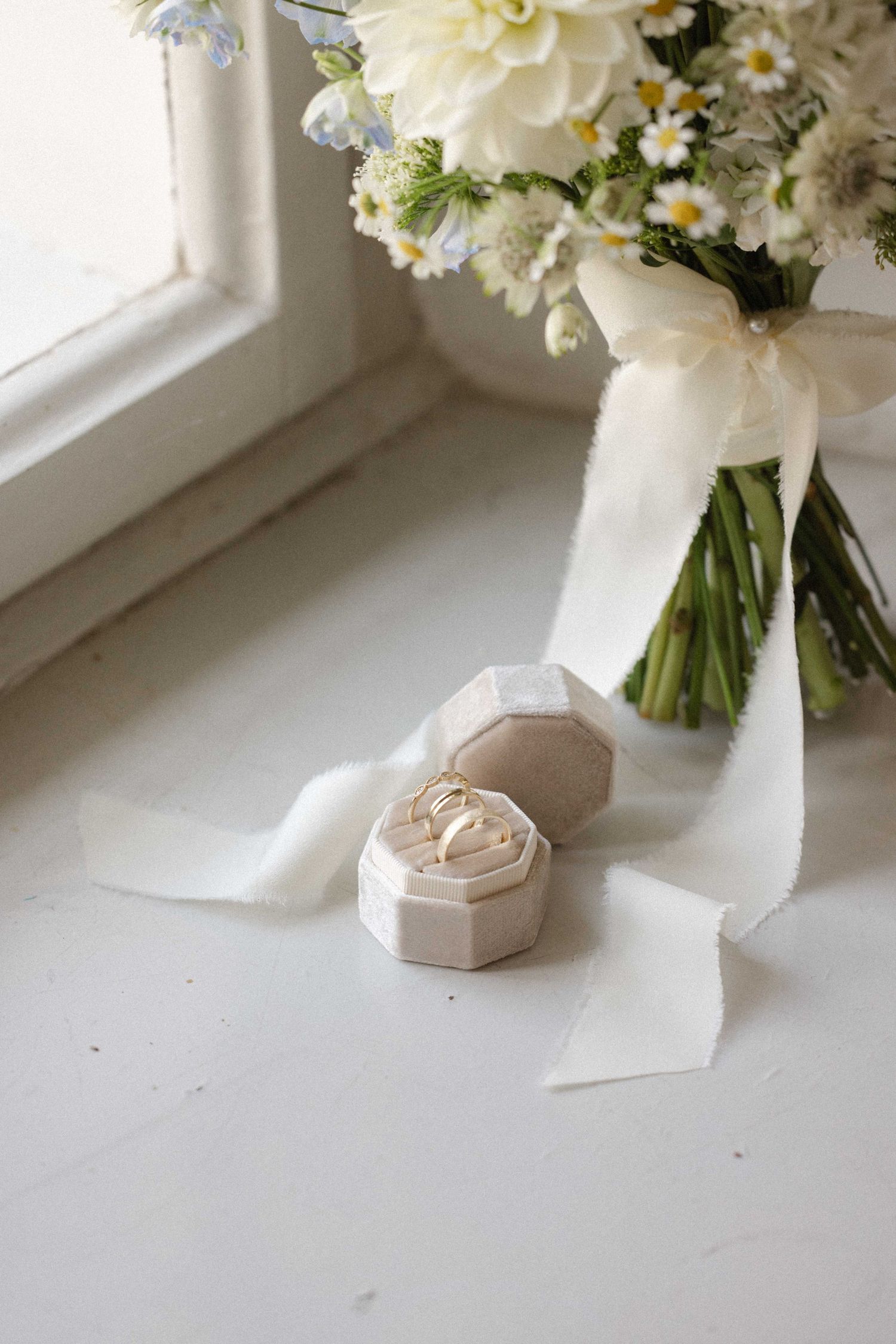 Wedding ring box with bouquet of white flowers and ribbon on windowsill for bridal detail shot.