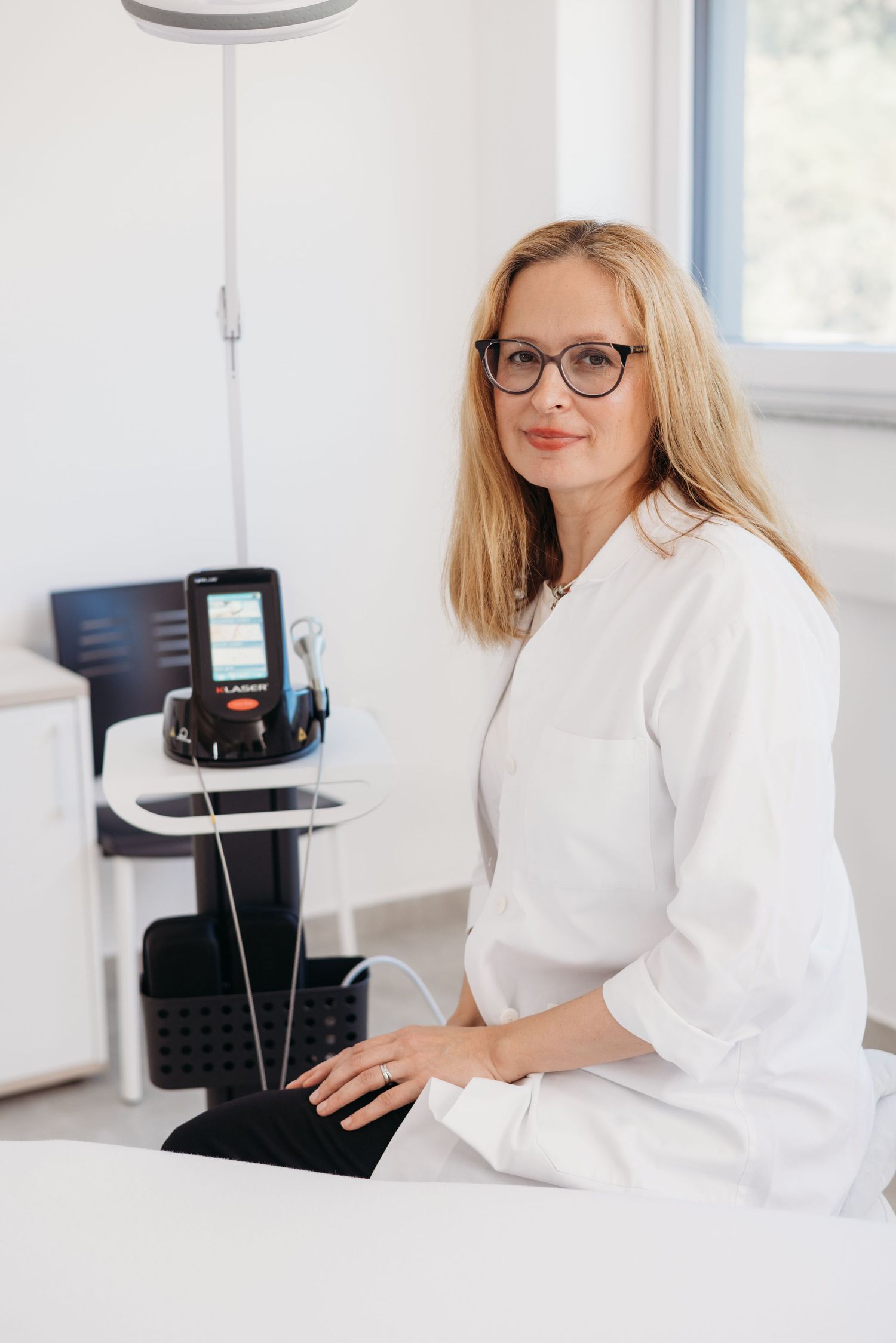 Female doctor in white coat and glasses sitting next to laser medical equipment in a bright clinical office.