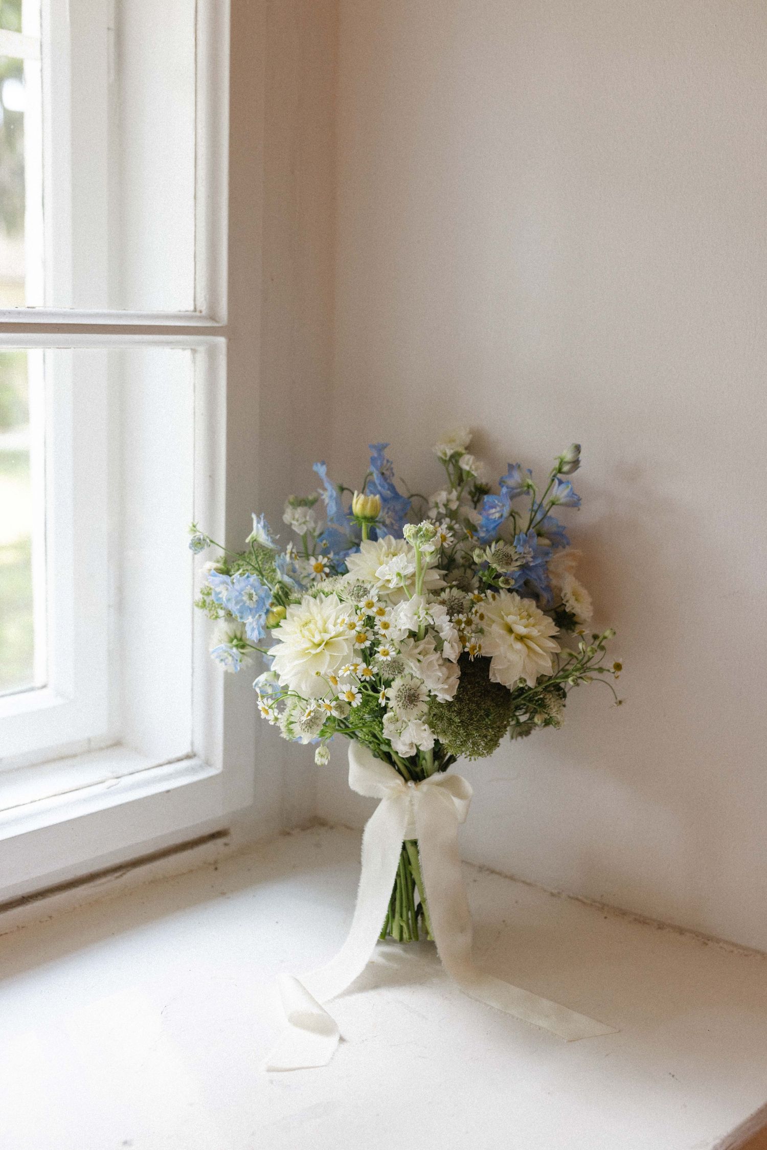 Elegant bridal bouquet of white and blue flowers tied with ribbon, placed by a sunlit window.